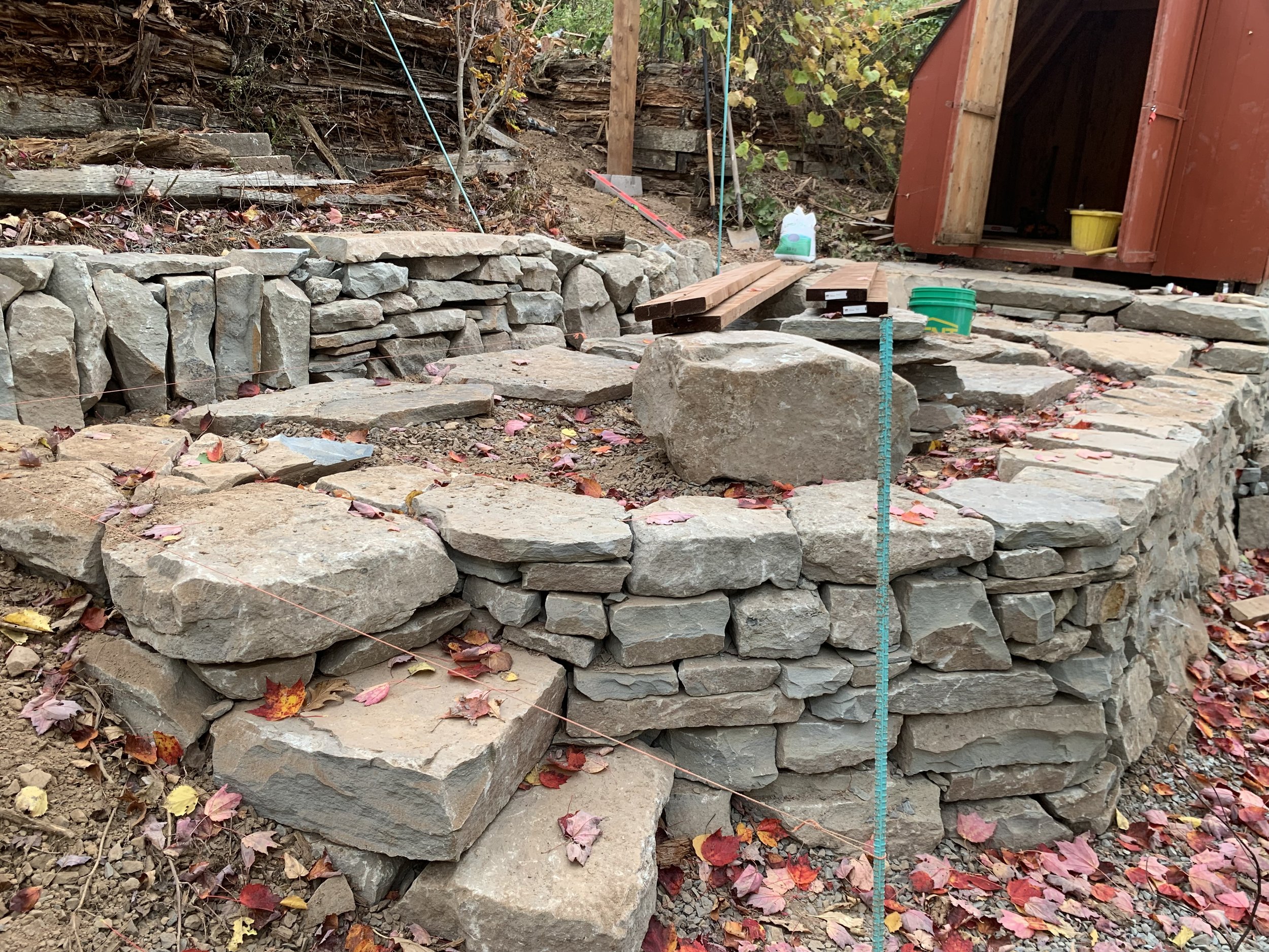 Stacked stone retaining wall under construction with construction tools and materials, surrounded by fallen autumn leaves.