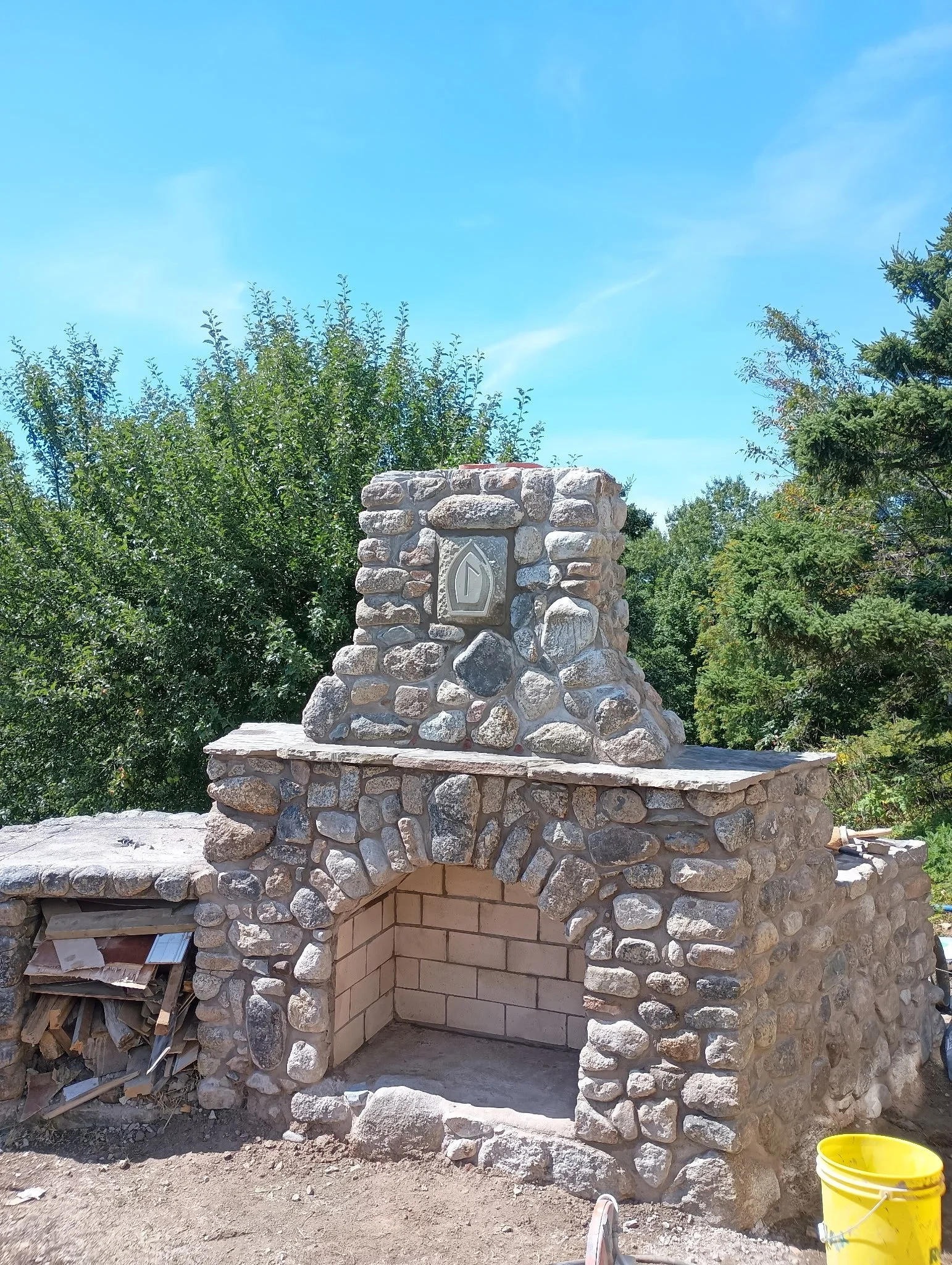 Stone outdoor fireplace under construction with a brick interior and a decorative stone chimney, set against green trees and blue sky.