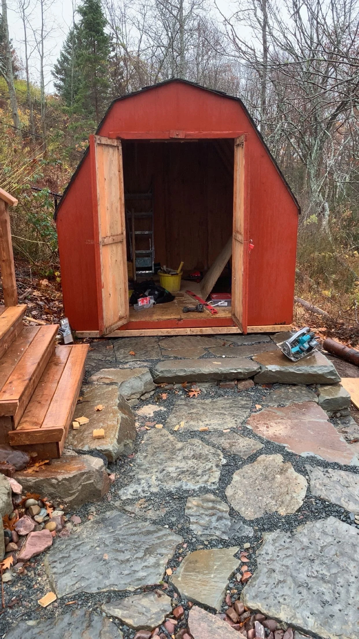 Small red wooden shed with open doors, tools and supplies inside, stone patio and steps leading up to it, surrounded by trees with autumn leaves.