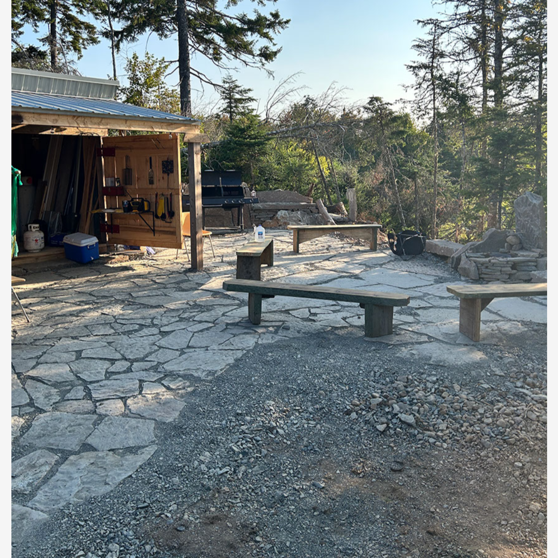Outdoor stone patio area with wooden benches, a shed with tools, a barbecue grill, and trees in the background.