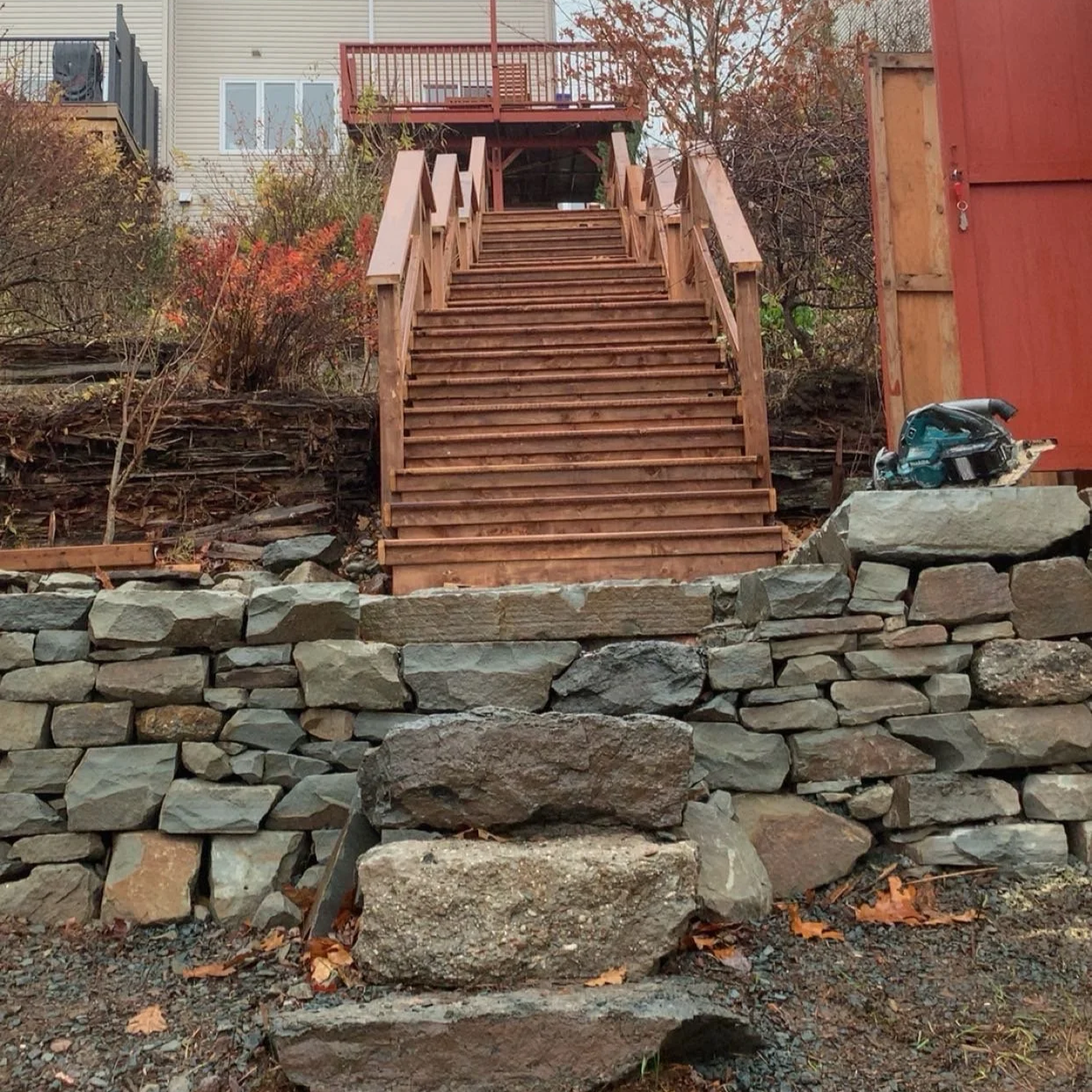 Wooden staircase leading up to a deck in a backyard, with a stone retaining wall at the base and construction tools nearby.