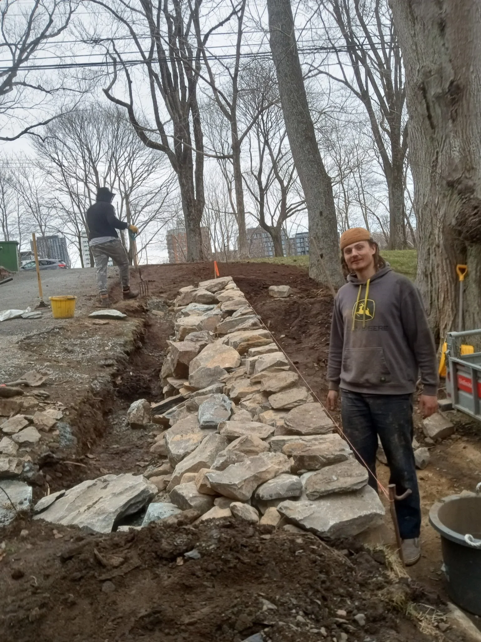 Two men constructing a stone wall on a hillside in a park with leafless trees and city buildings in the background. One man is standing near the stone wall, while the other is working on the slope.