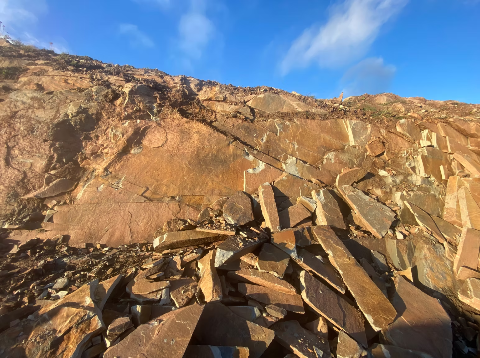 Close-up of a rocky Quarry-face with large and small brown and tan rocks, some cracks, under a blue sky with a few clouds.