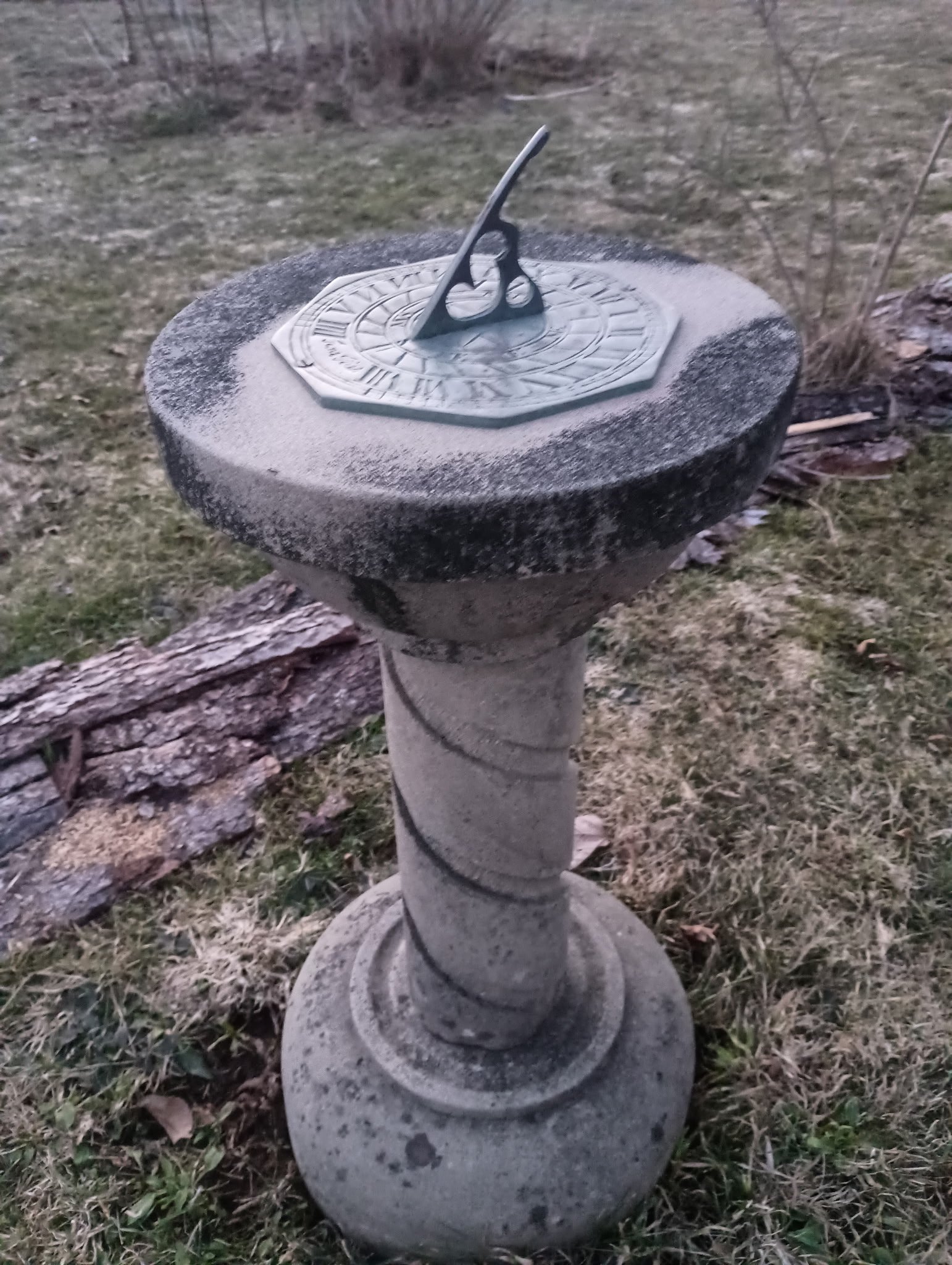 A sundial on a stone pedestal outdoors, with grass and a fallen log nearby.