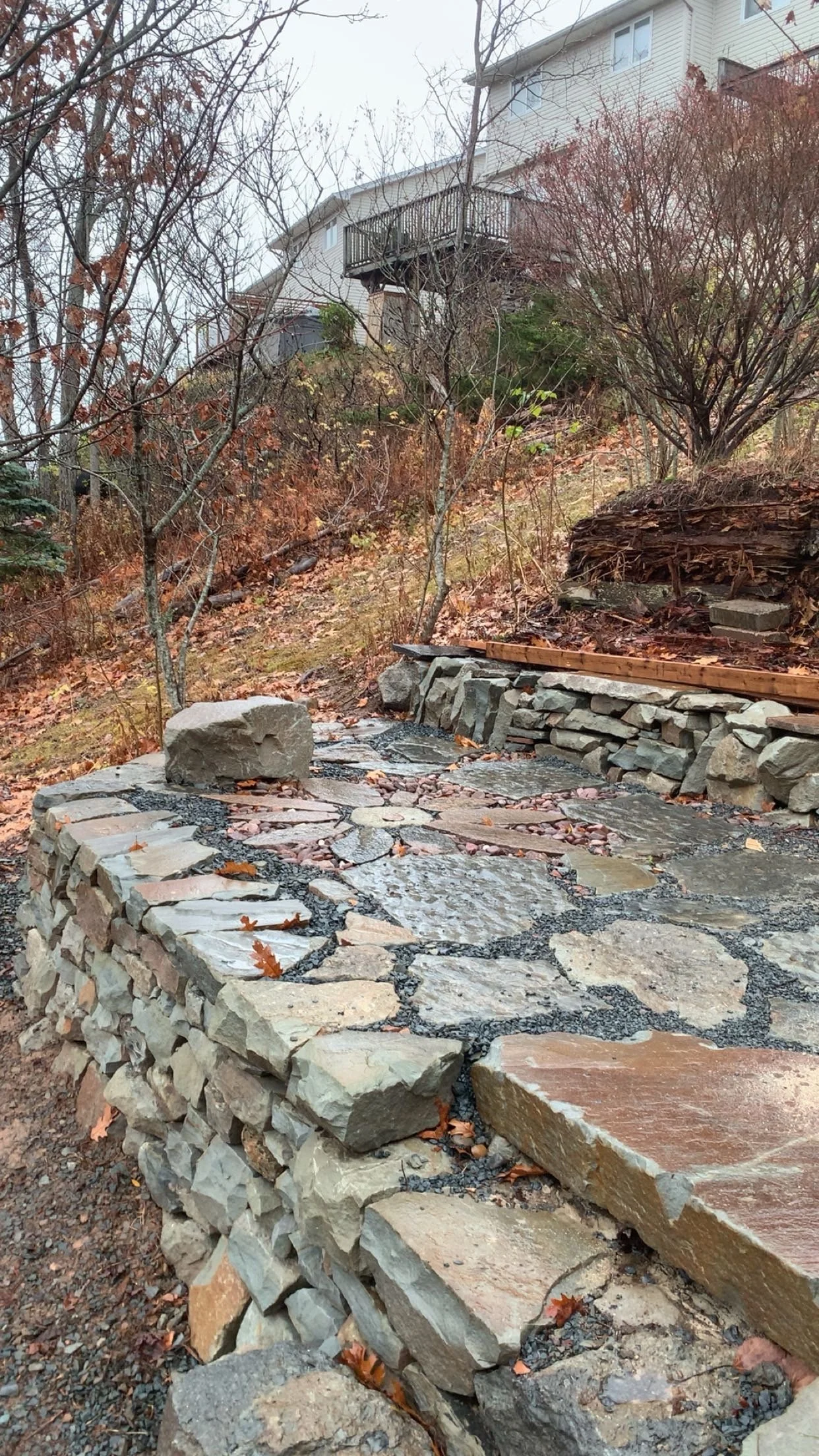 A stone pathway on a hillside with autumn leaves and sparse trees, with houses and a wooden balcony in the background.