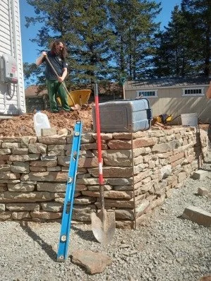 Person standing above a stone retaining wall with a shovel hanging on a level tool and a blue level nearby, outdoors with trees and a building in the background.