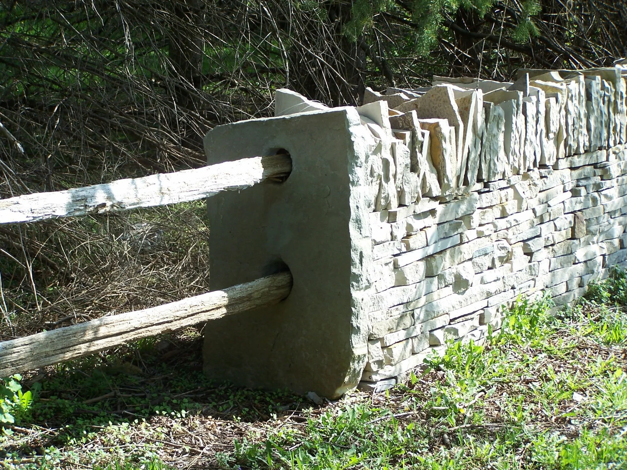 A stone and wood fence in a natural outdoor setting, with green grass, small plants, and dense bushes in the background.