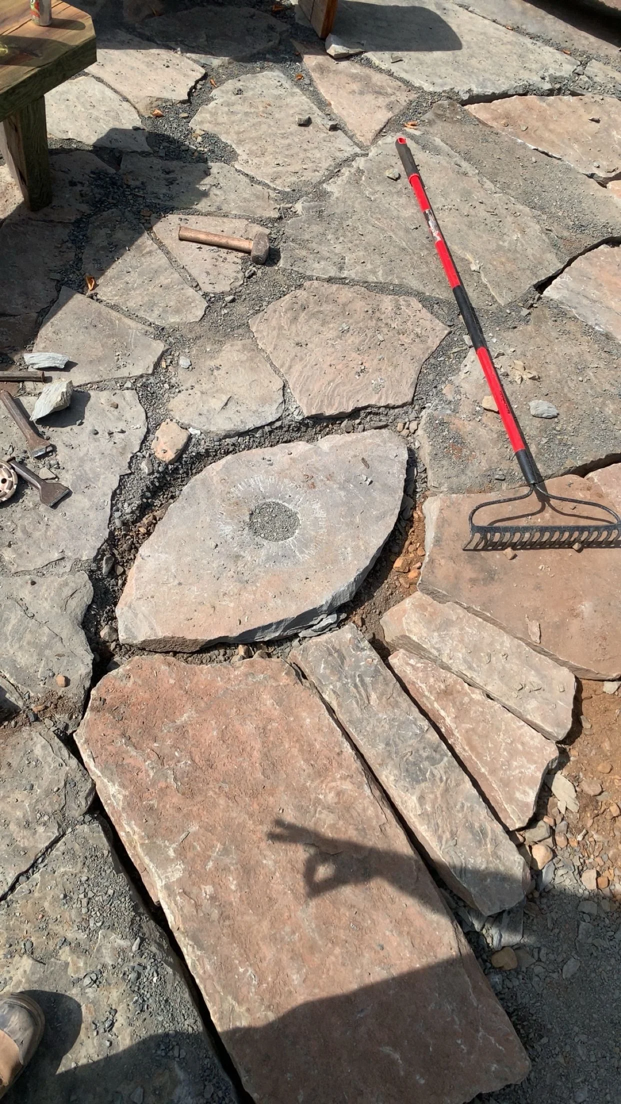 Unfinished stone patio with various large flat stones and a carved stone eye in the center, some tools like a rake and a hammer, and a shadow of a person taking the photo.