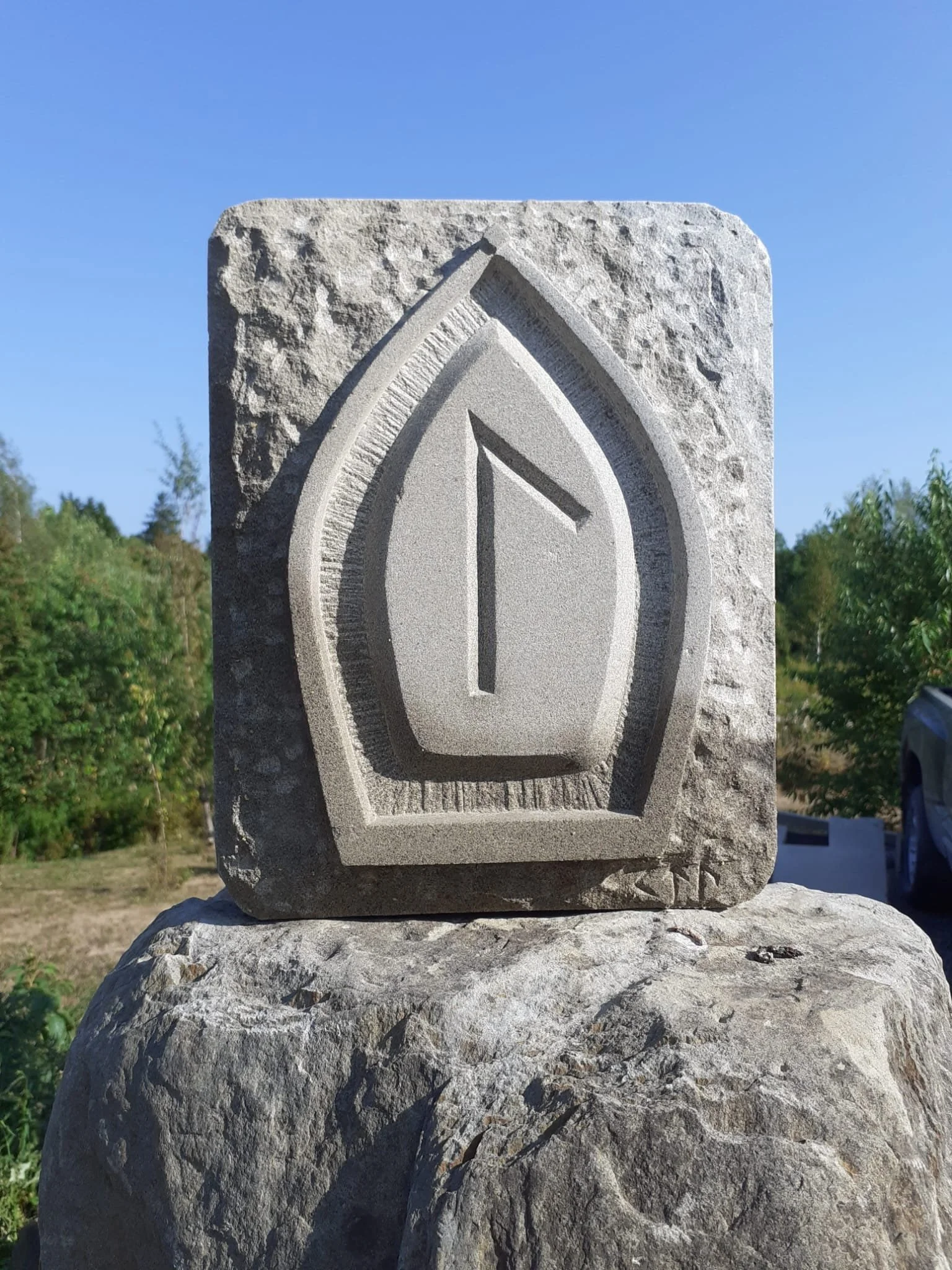 A carved stone monument with a geometric design resembling a stylized row boat, featuring a central Lagu rune, situated outdoors on a large rock with trees and a clear blue sky in the background.