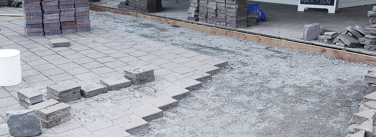 A construction site where brick pavers are being laid on the ground. Some bricks are stacked, and others are already placed. There is a white bucket on the left and a blue object on the raised platform in the background.