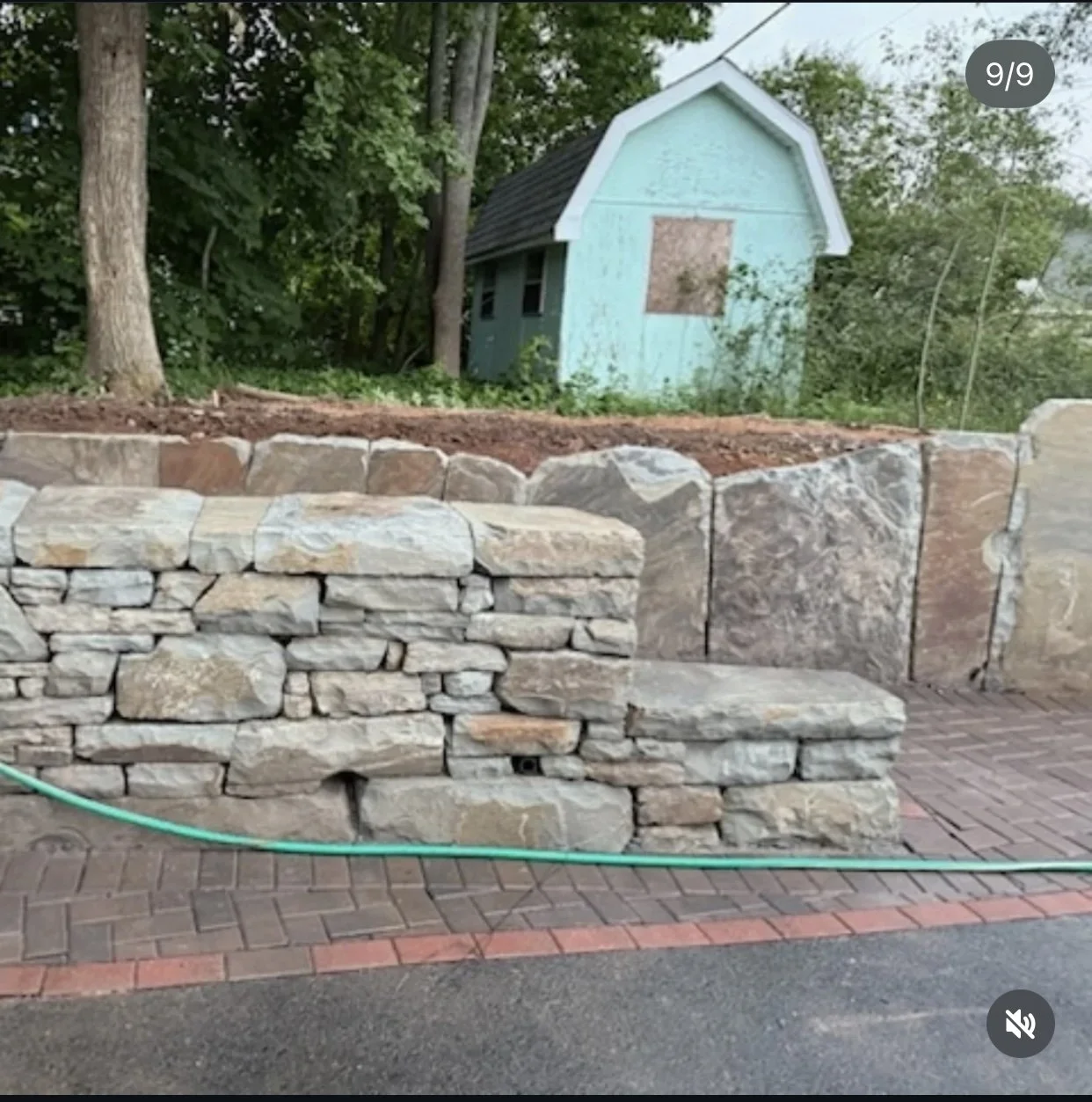 A stone retaining wall with a brick and stone pattern, with a garden hose in front, and a small blue shed in the background among trees.