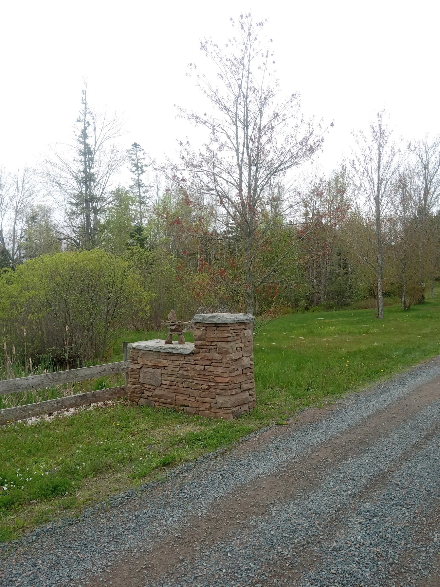 A stone structure with a small sculpture on top is located next to a gravel road, with green grass and trees in the background.