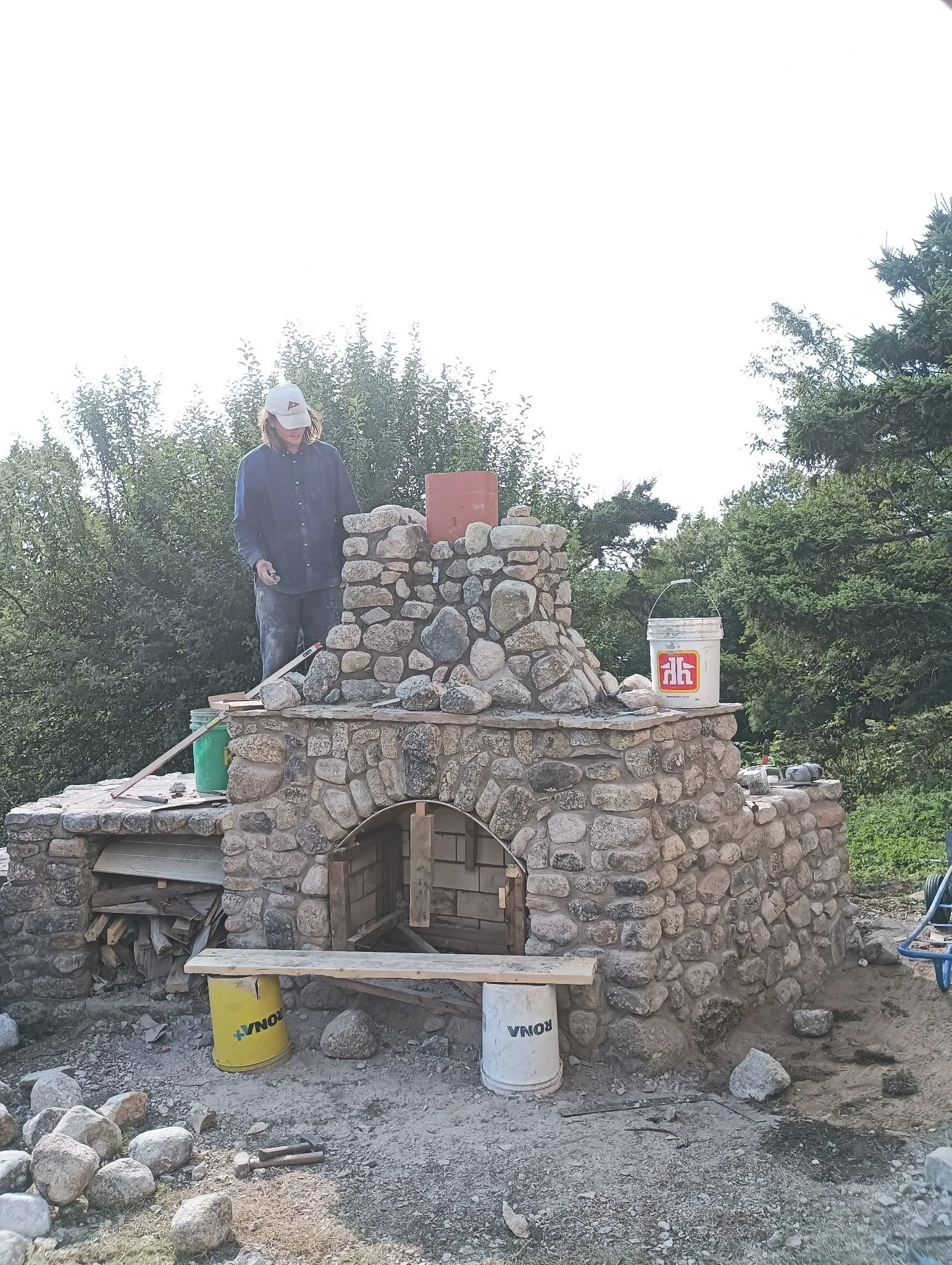 Person constructing a stone oven or fireplace outdoors, surrounded by trees and construction materials.