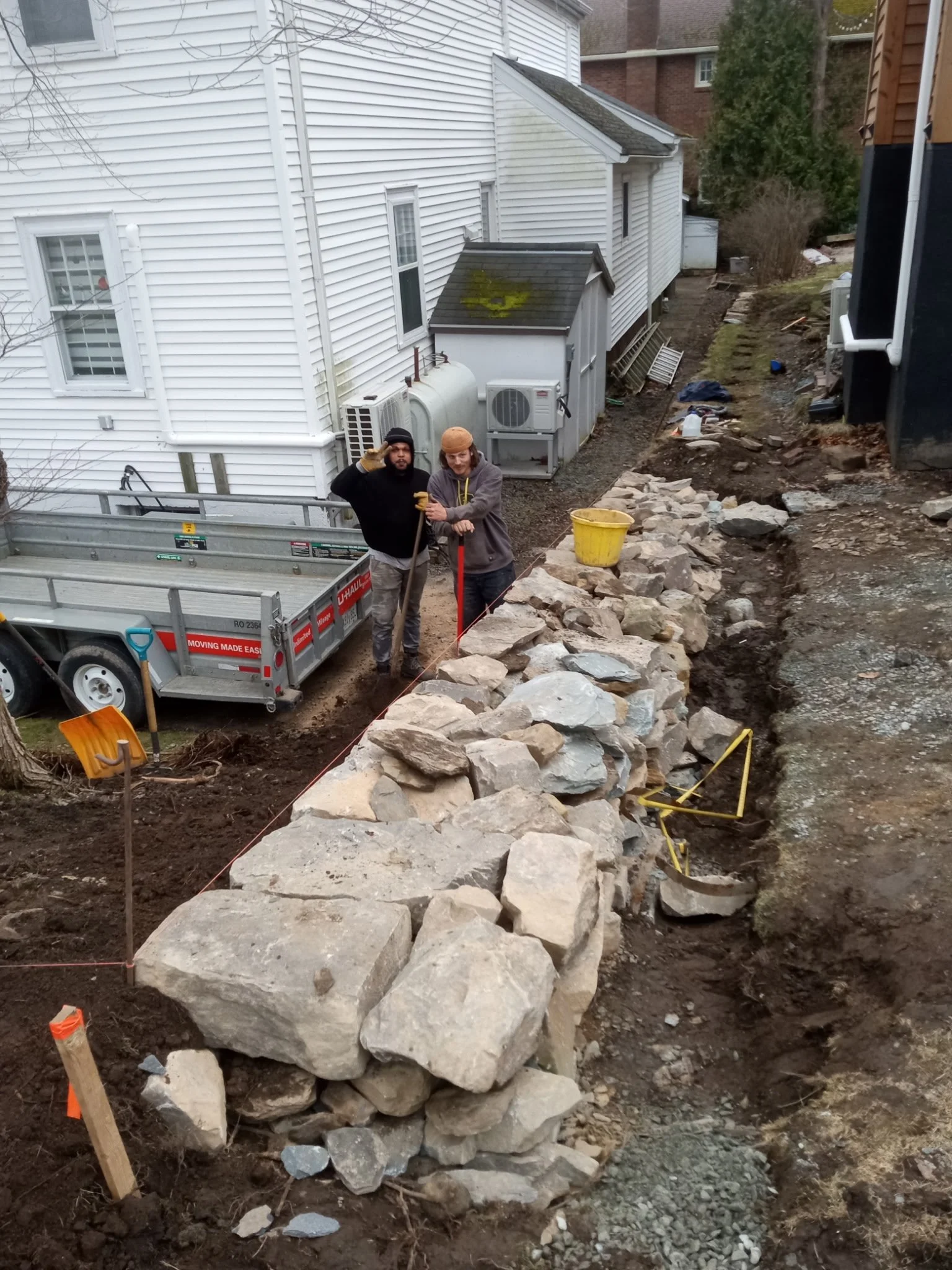 Two construction workers standing on a stone wall under construction in a backyard, with a trailer and house in the background.