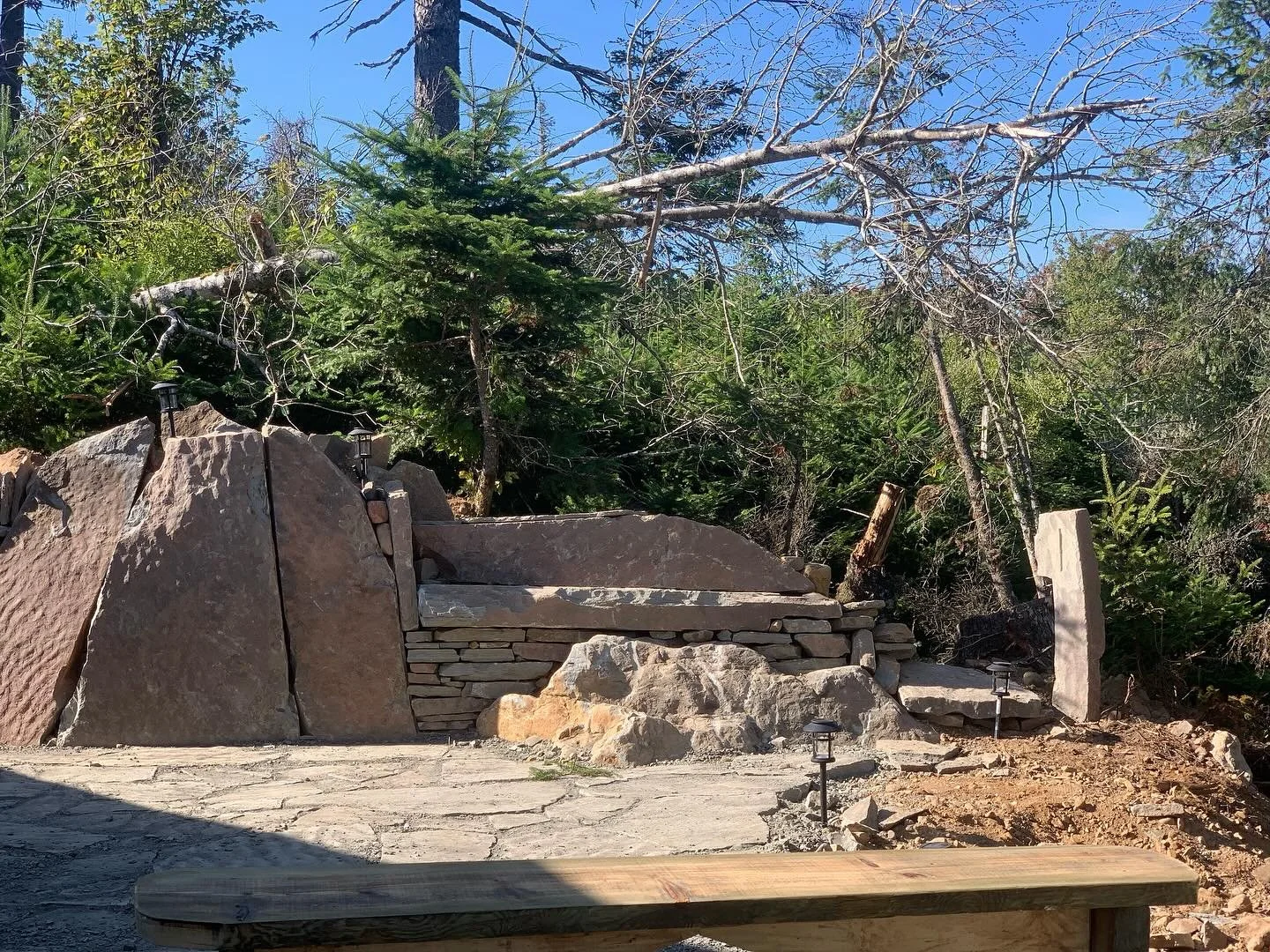 A stone wall and steps leading up to a natural outdoor area with trees and greenery, with small pathway lanterns along the ground.