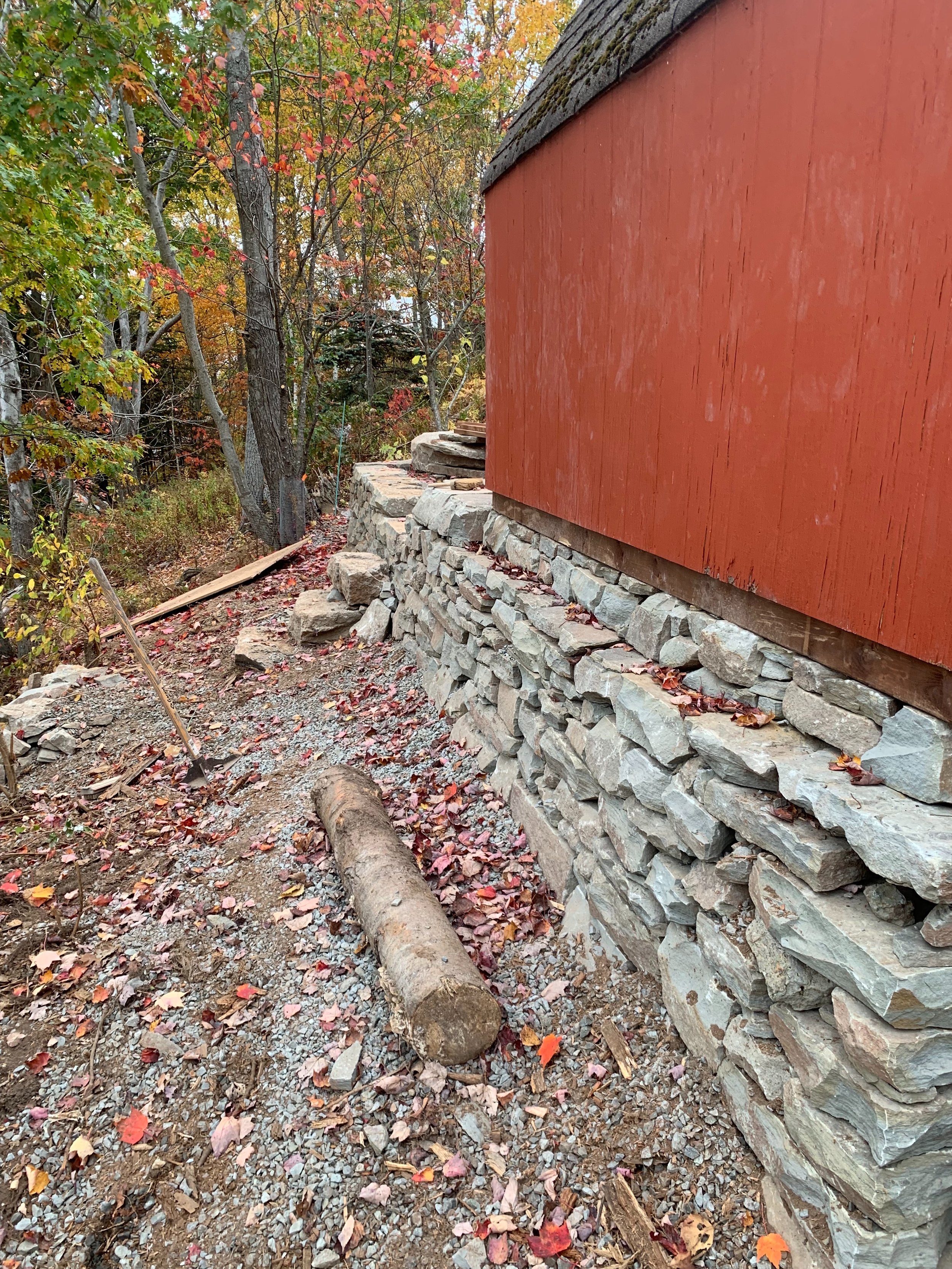 Construction site with a stone foundation next to a red wooden building, autumn leaves scattered on the ground, and trees with fall foliage in the background.