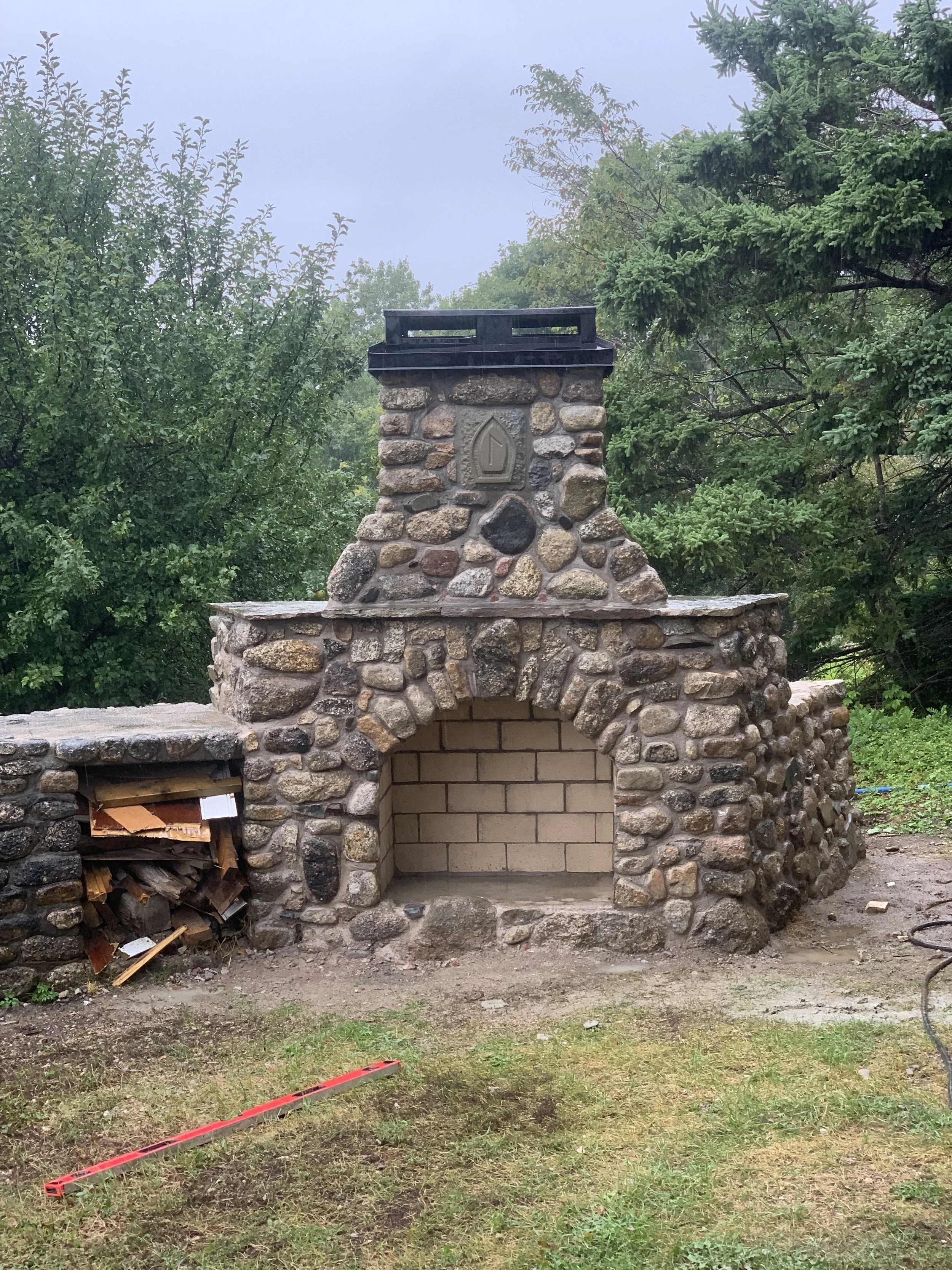Stone outdoor fireplace with a built-in oven on the left and surrounded by green trees.