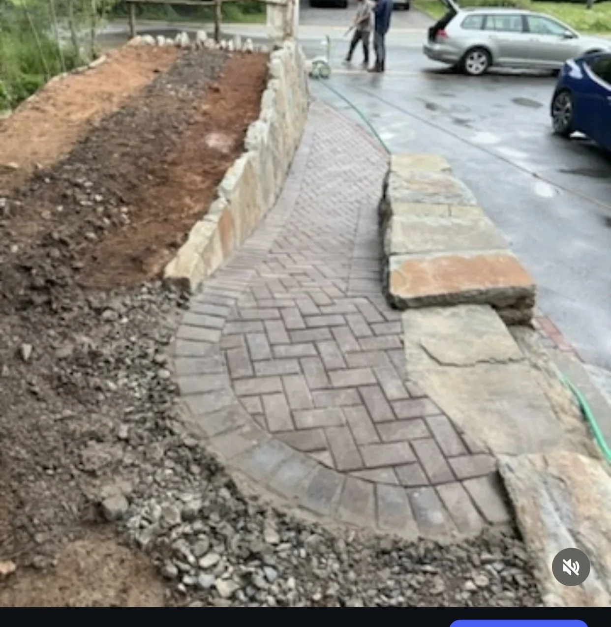 A newly constructed brick sidewalk with a curved pattern, bordered by large stones, next to a parking lot with cars and people in the background.
