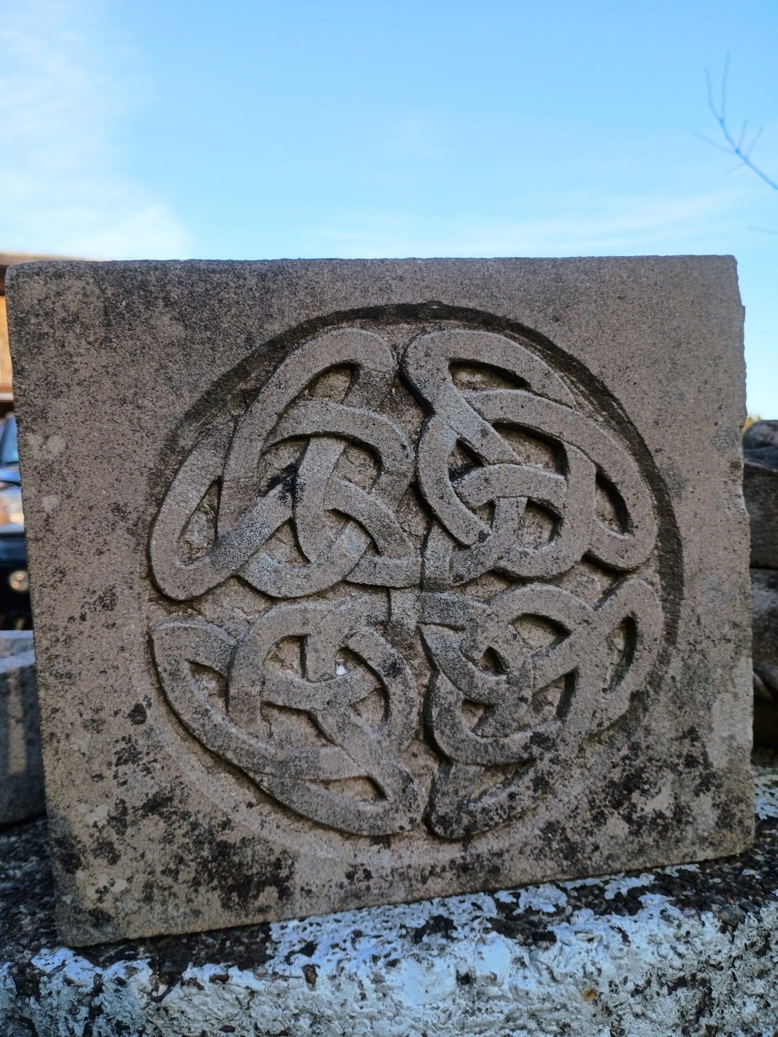A square stone with intricate Celtic knot design carved into it, placed outdoors against a background of blue sky.