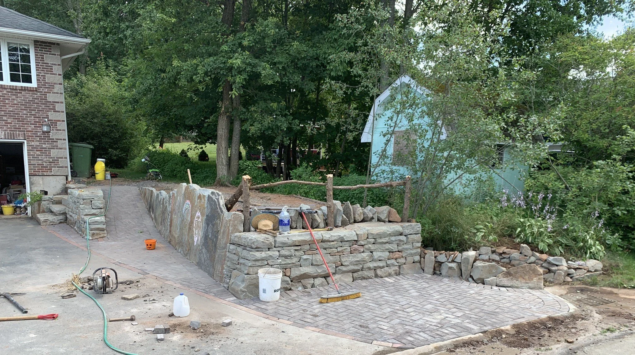 Landscaping in progress with a stone retaining wall under construction, brick paving, and various tools and materials around a residential yard.