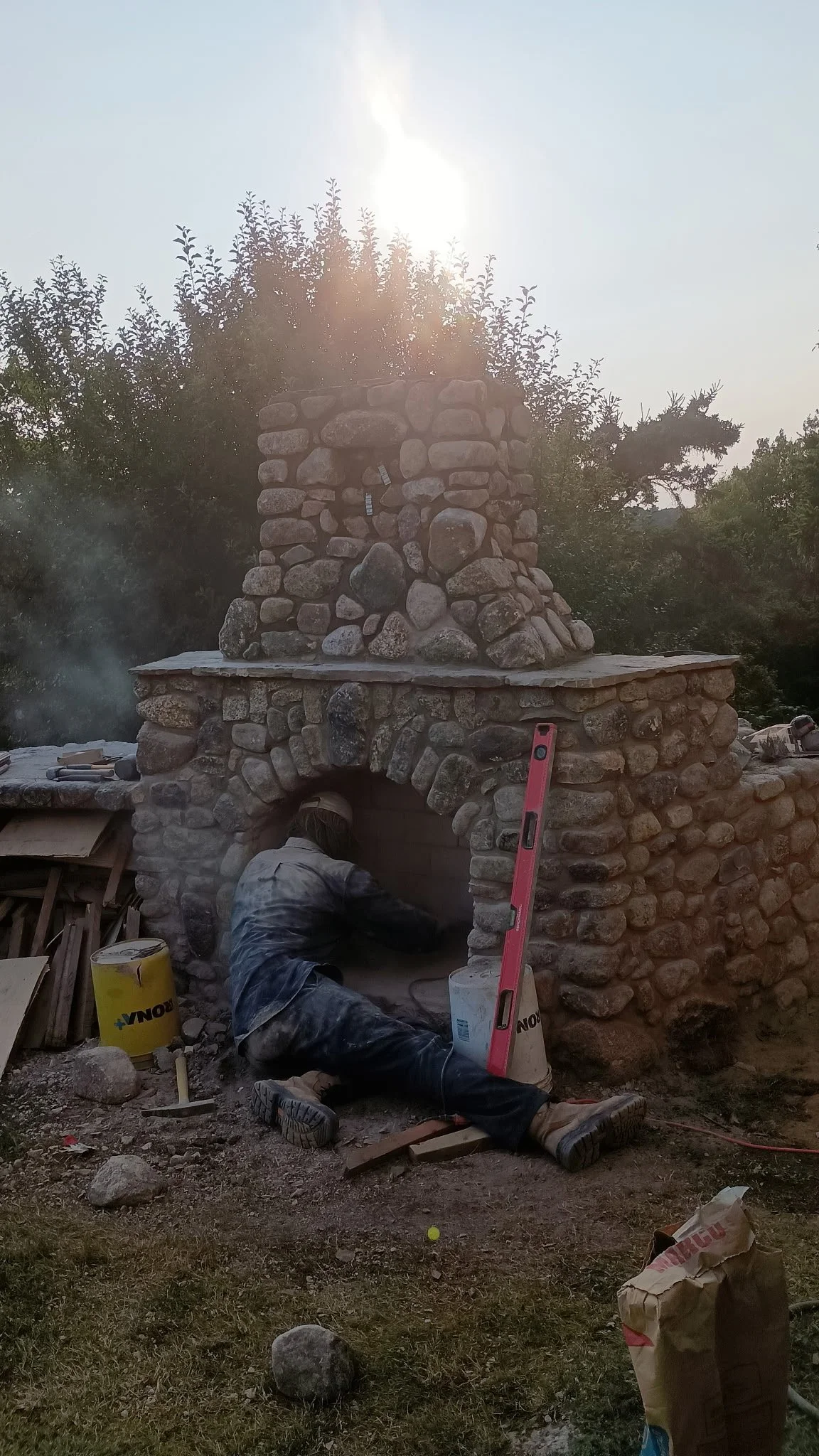 A person working on constructing a stone outdoor fireplace or oven. They are kneeling inside the structure, surrounded by construction tools and materials, with a tall stone chimney and a work level visible. The setting is outdoors in a yard with tre