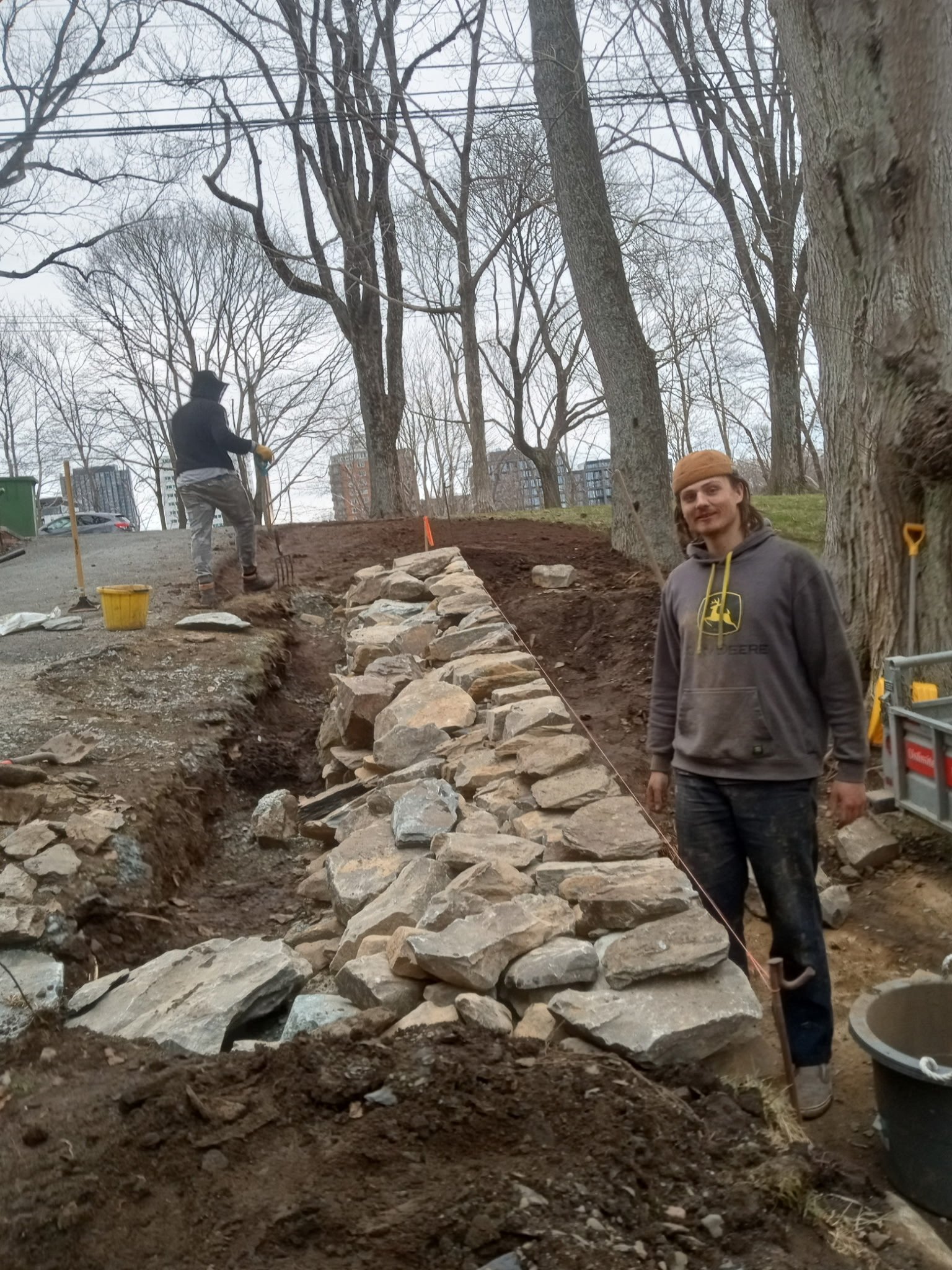 Two men working outdoors on a landscaping project. One man is standing on the ground and smiling at the camera, wearing a brown beanie and a gray hoodie. The other man is in the background, working on a stone wall or pathway, wearing gloves, a black 
