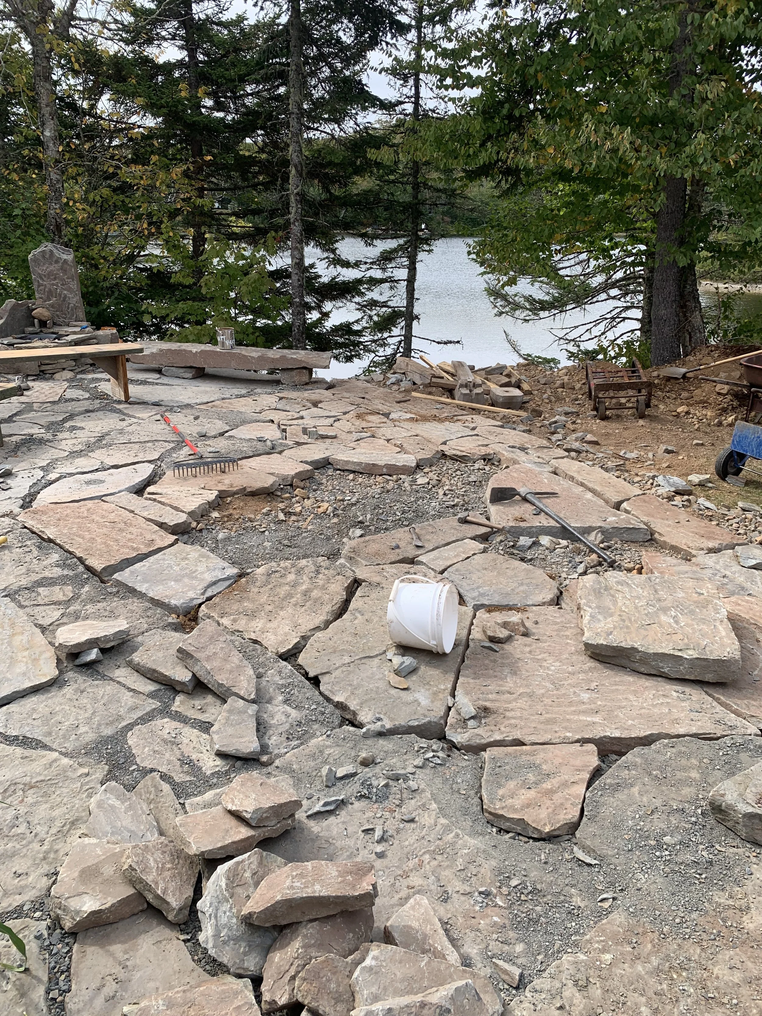 Under construction outdoor patio with large flat stones, construction tools, and a white bucket, surrounded by trees and near a body of water.