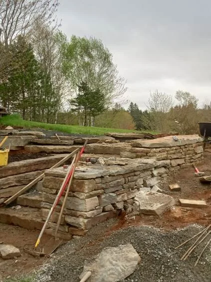 A stone wall and stairs under construction with tools and materials around it, set outdoors with trees and cloudy sky in the background.