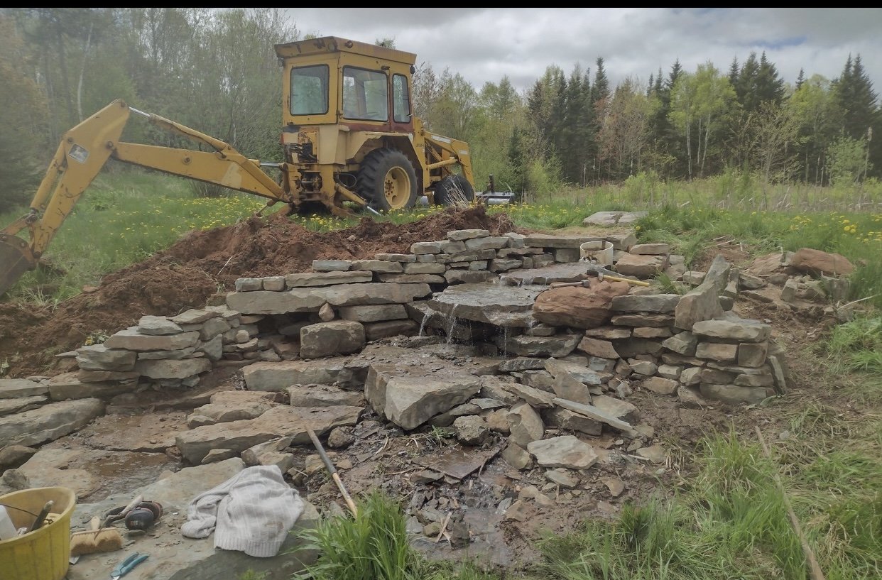 Construction site with a yellow backhoe and a partially built stone wall over a small water stream in a grassy field with trees in the background.