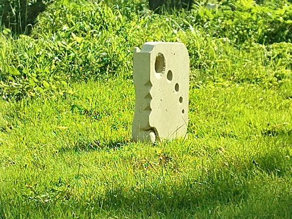 A Stone geometric sculpture with multiple holes and cutouts standing on green grass, surrounded by bushes.