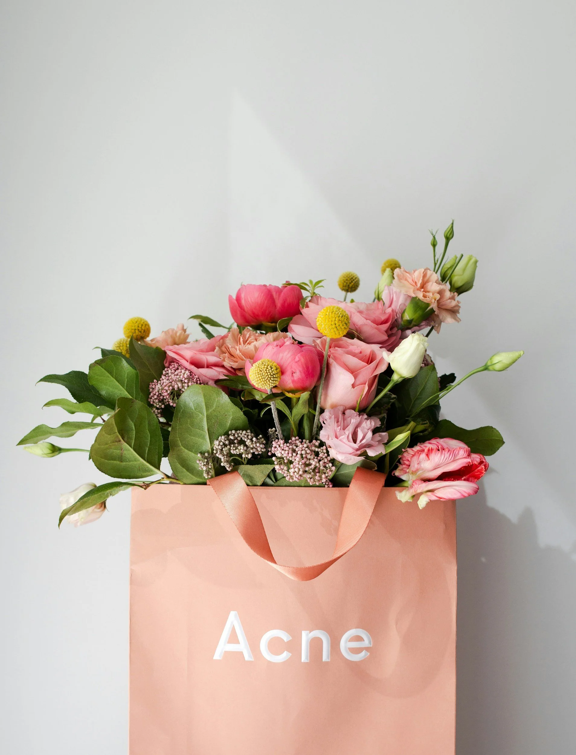 A bouquet of pink, white, and yellow flowers in a pink paper gift bag with the word 'Acne' printed on it.