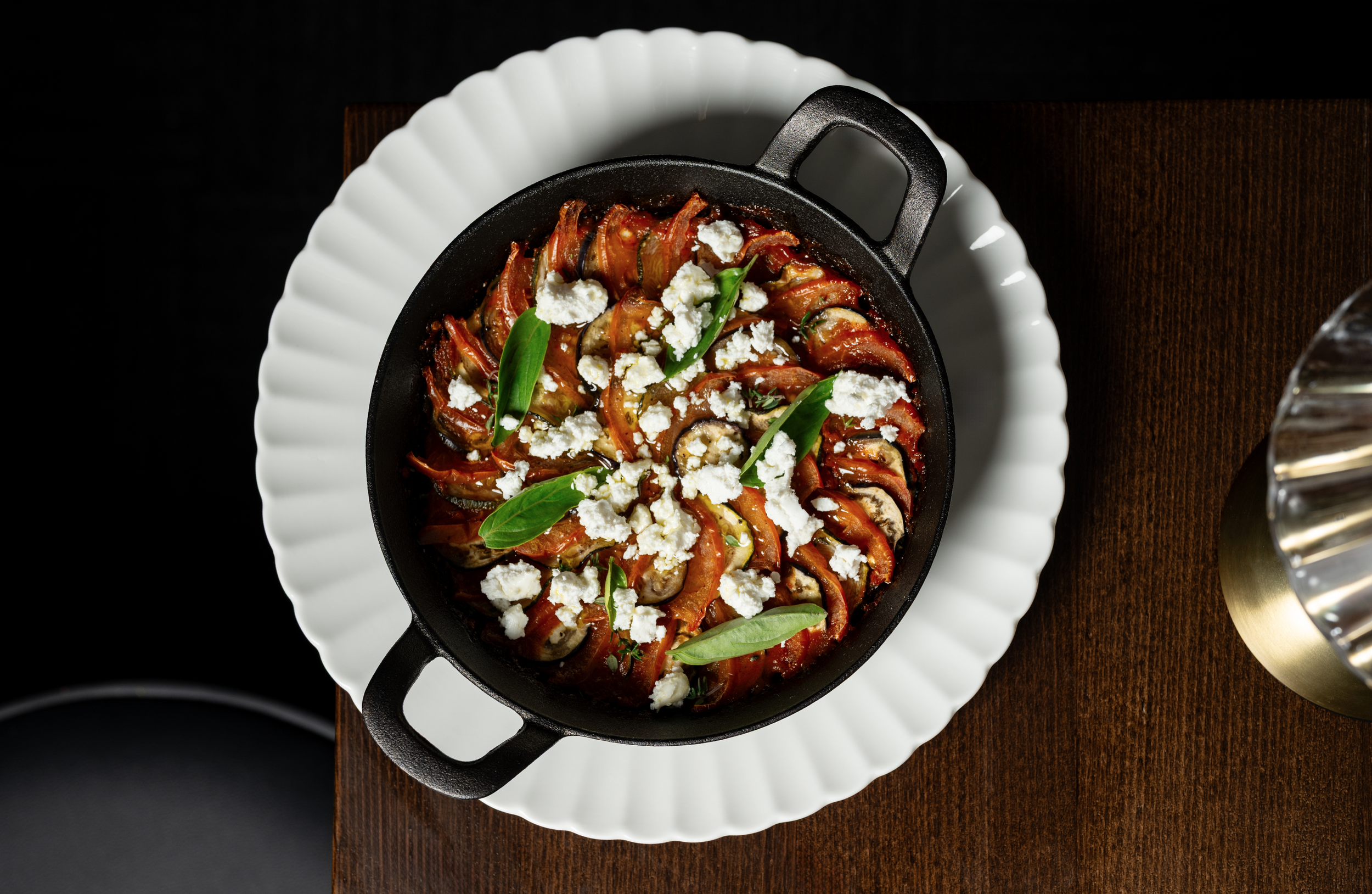 A skillet of roasted vegetables topped with crumbled cheese and fresh basil leaves, served on a white plate on a wooden table.