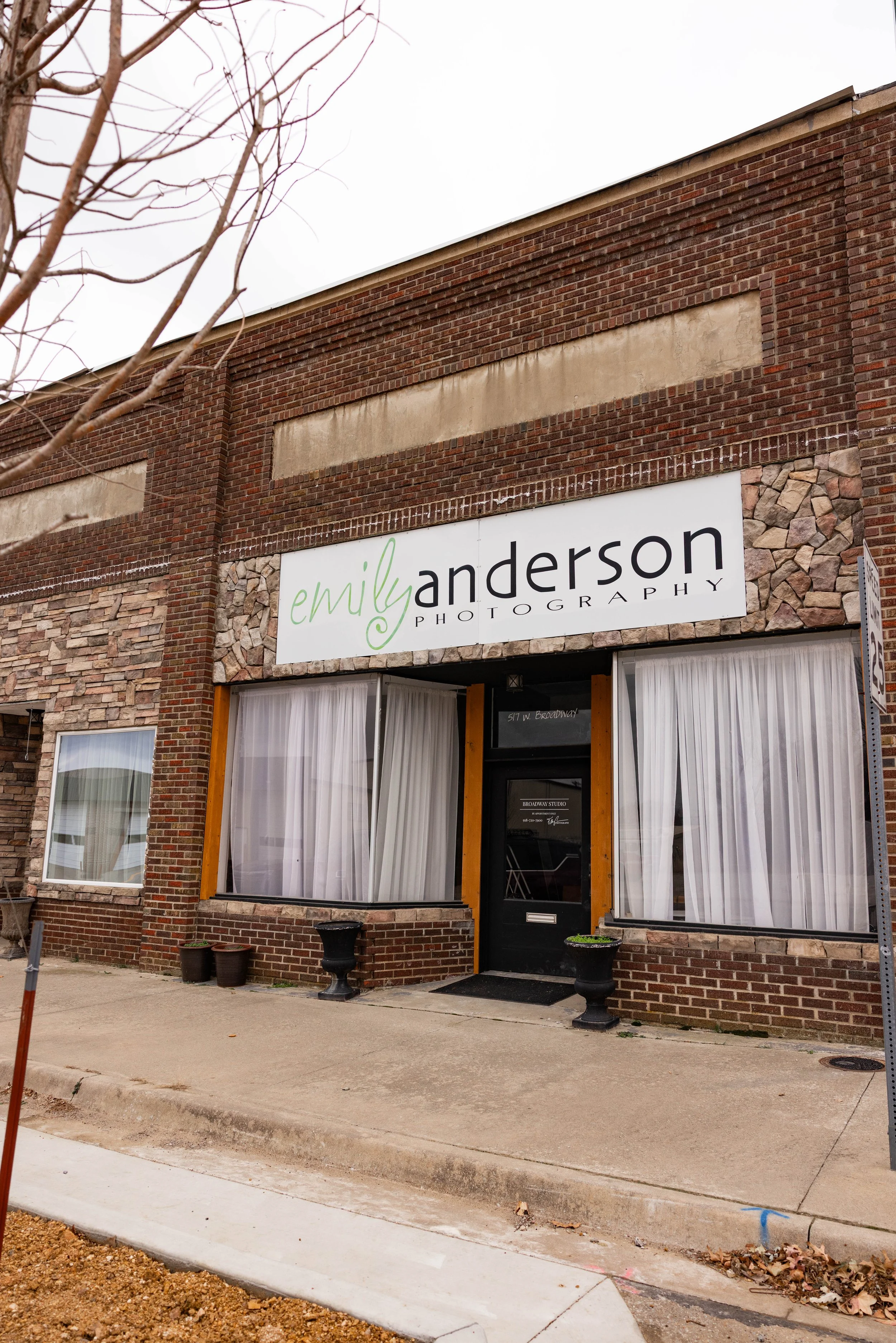 storefront with a sign reading 'emil anderson photography' above the entrance. The building has red brick walls, large windows with white curtains, and potted plants outside.