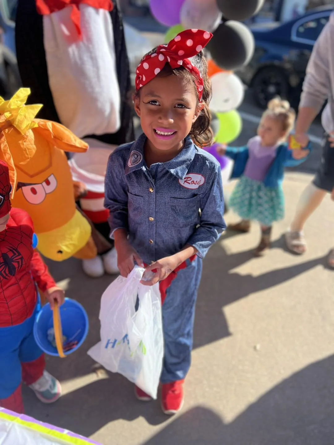 A young girl wearing a red polka dot headband and denim jumpsuit smiling at a fall festival, holding a white bag, with children and Halloween-themed decorations in the background.