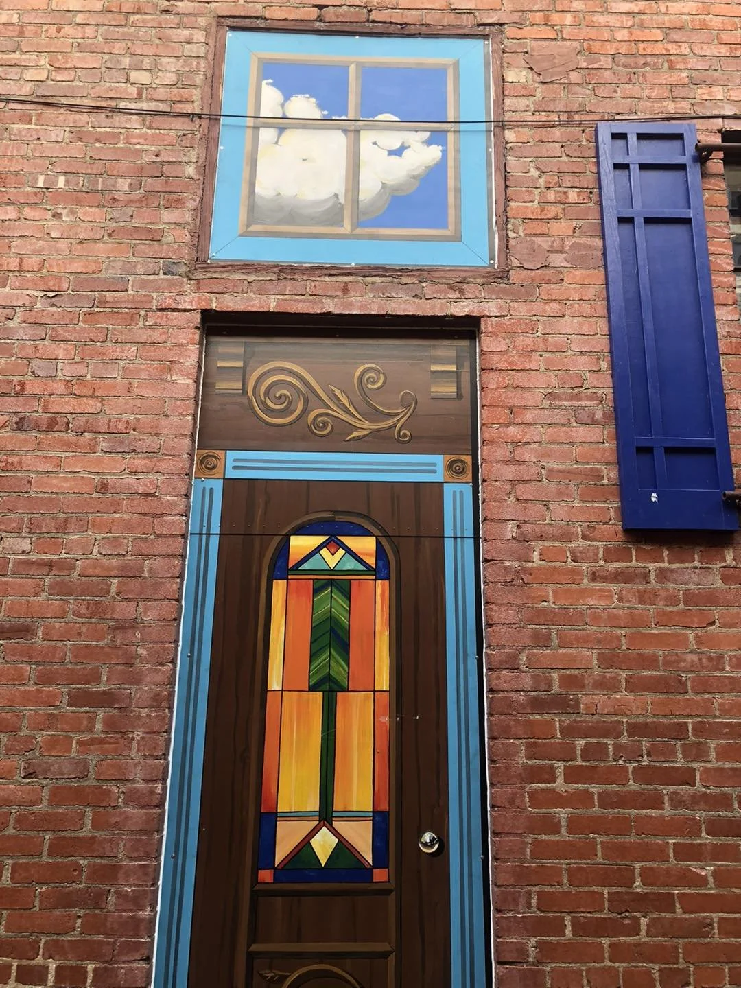 Brick building with a door featuring a stained glass window and a window with a painted sky and clouds.