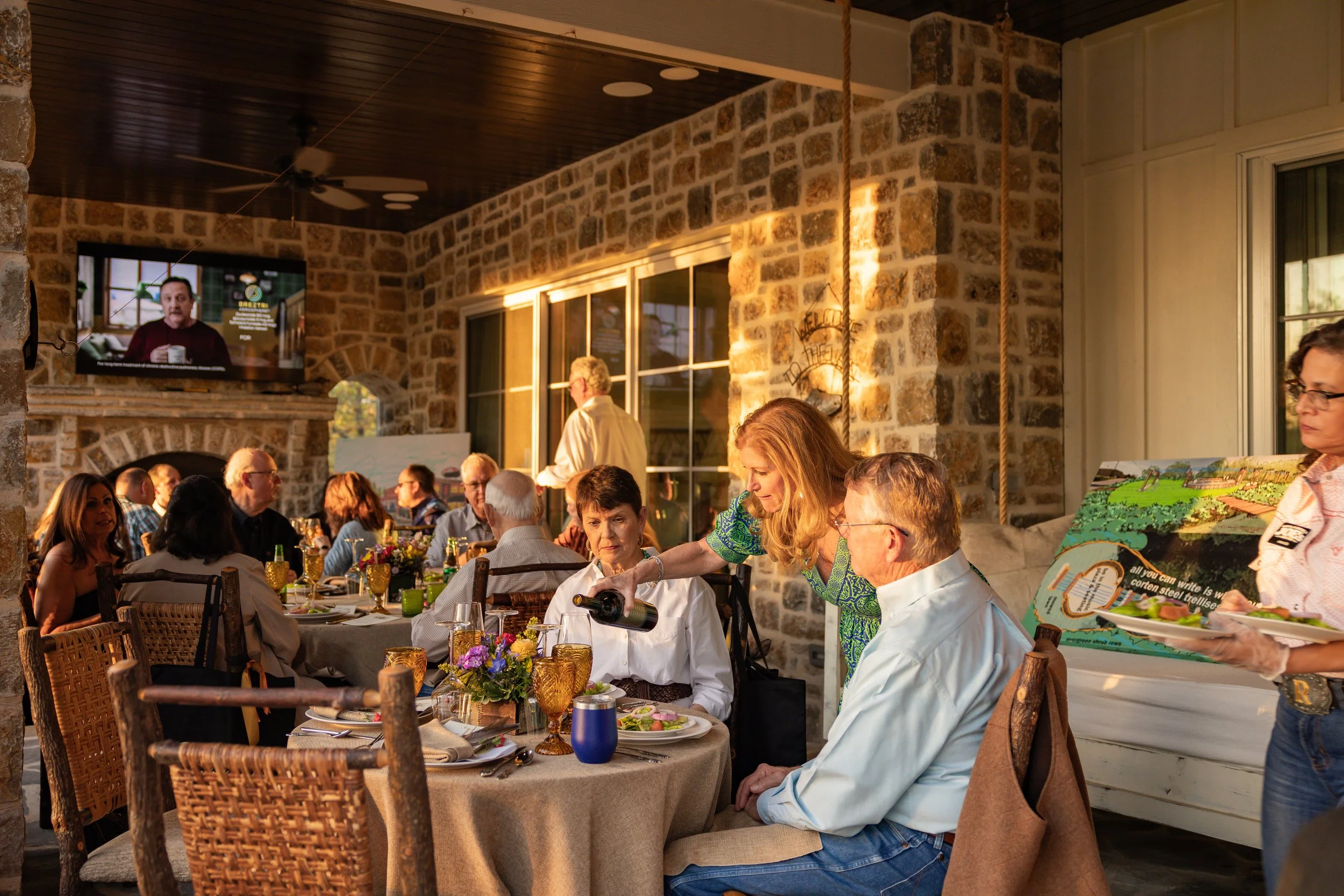 People gathered at a dinner party in a rustic, stone-walled room with a fireplace and a mounted television.