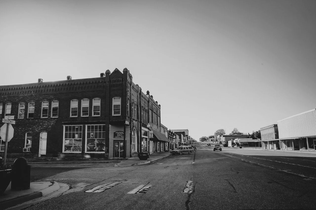 Empty street in a small town with buildings, parked cars, and a clear sky.