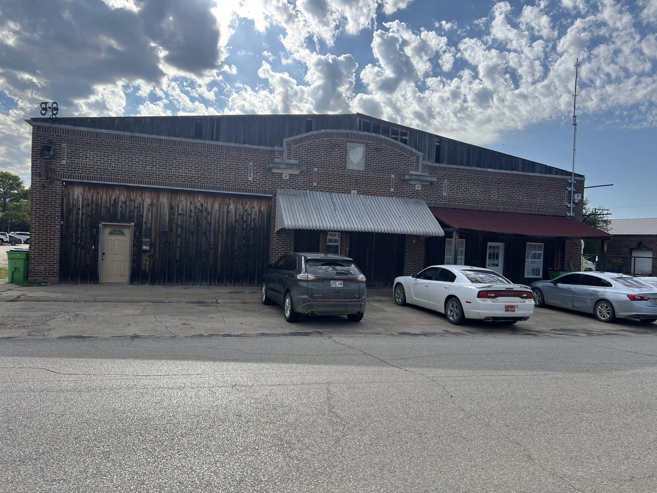 A brick building with a mix of materials, including dark wood and metal, with cars parked in front. The sky is partly cloudy.