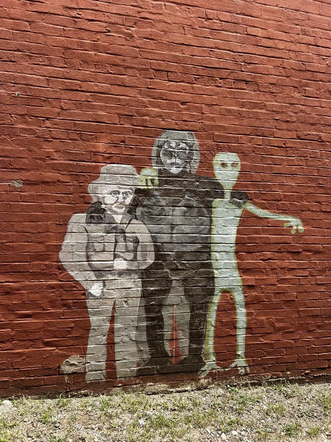 Street art graffiti of three humanoid characters painted on a red brick wall. The left character is a person with glasses, a hat, and a jacket. The middle character has a round head with a star on its face and is dressed in dark clothing. The right c