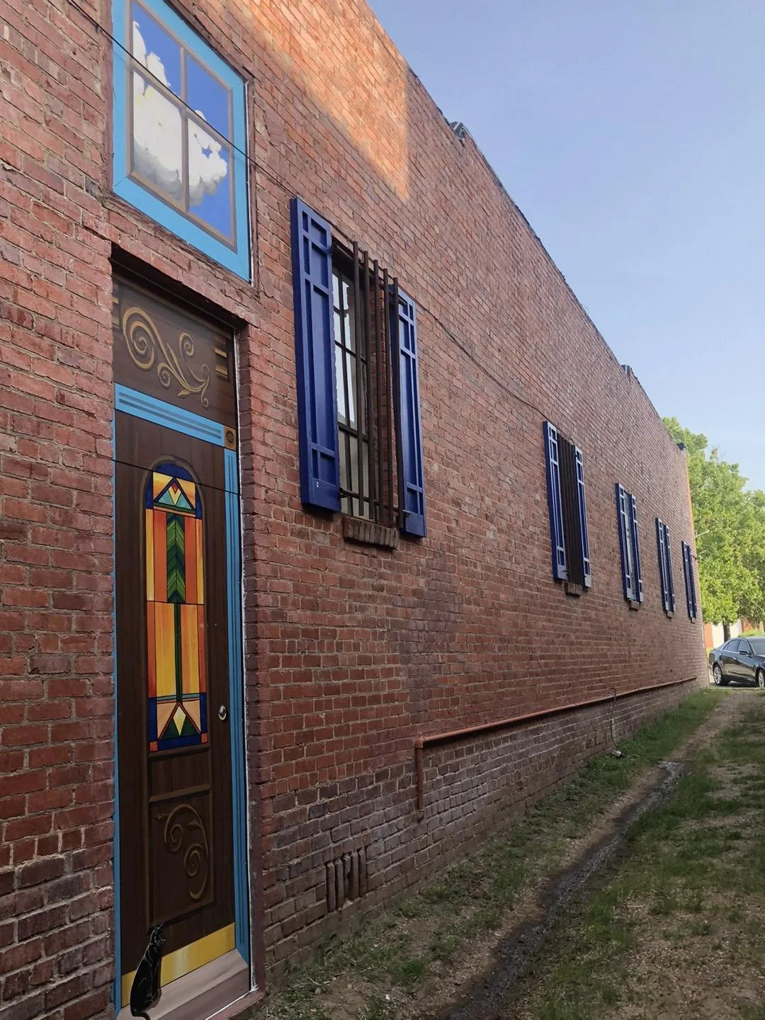 Brick building with colorful stained glass door, four tall blue windows with shutters, reflection of clouds in the top window, and trees in the background.