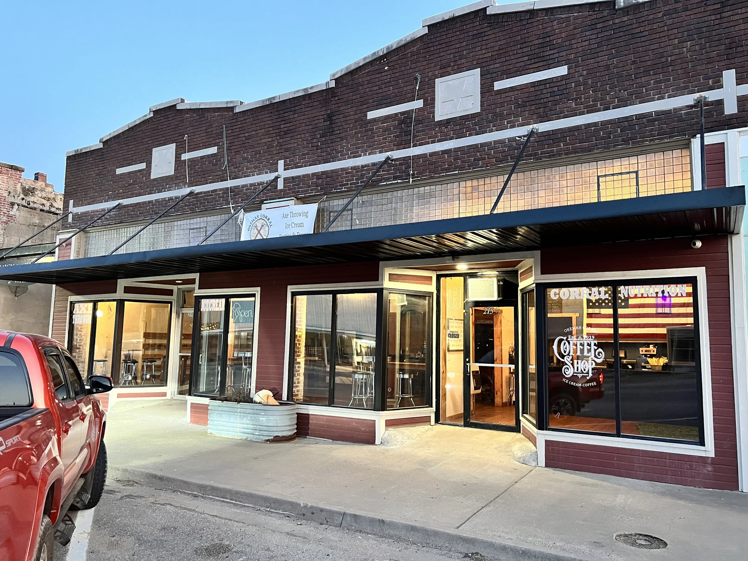 Exterior of a coffee shop with large glass windows and a glass door, parked red truck in front, and a brick building behind with signage indicating tea and ice cream offerings.