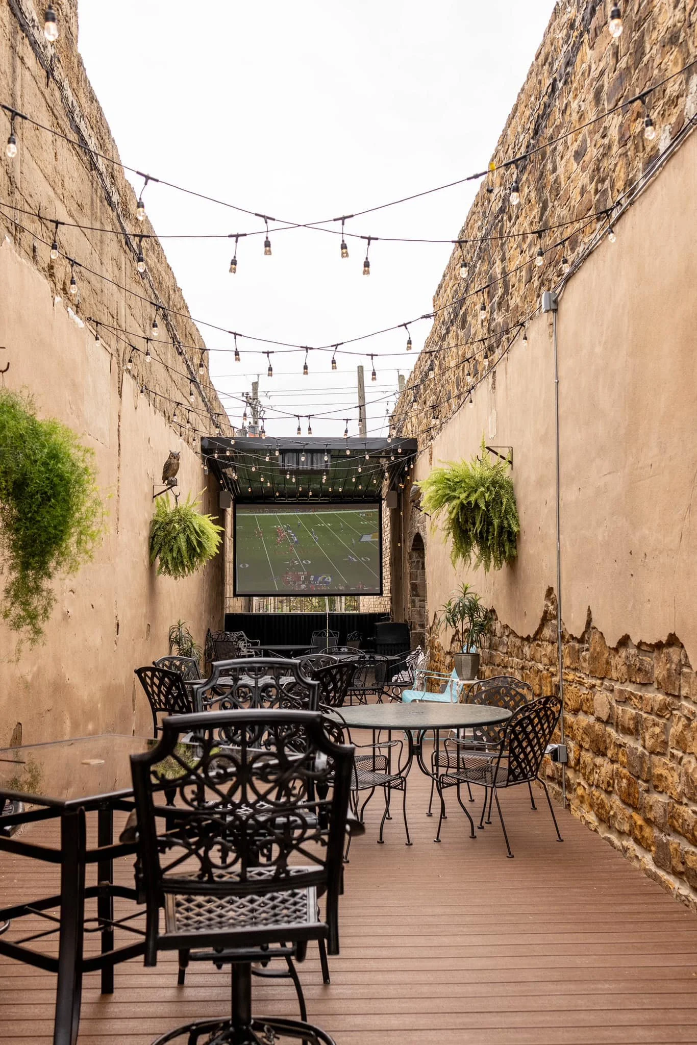 An outdoor patio with tables and chairs, hanging string lights, potted plants, a large screen showing a football game, and brick and stucco walls.