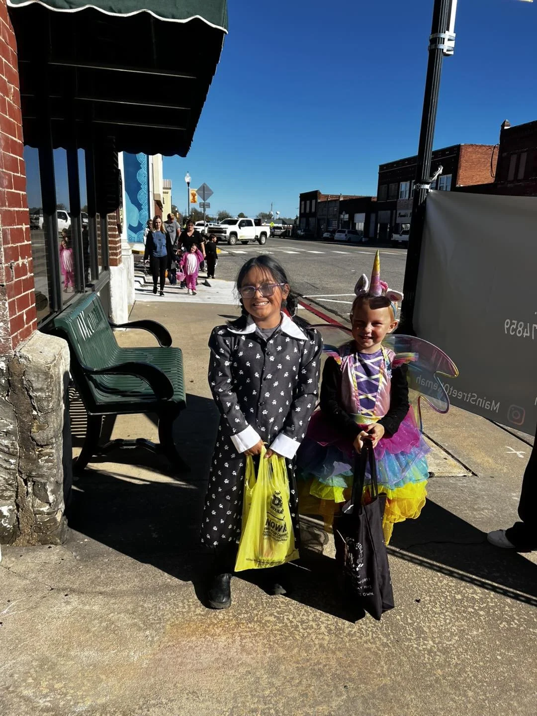 Two young girls dressed in costumes, one as a rainbow fairy and the other as a black and white witch, standing on a sidewalk and smiling at the camera during a festive event on a sunny day.