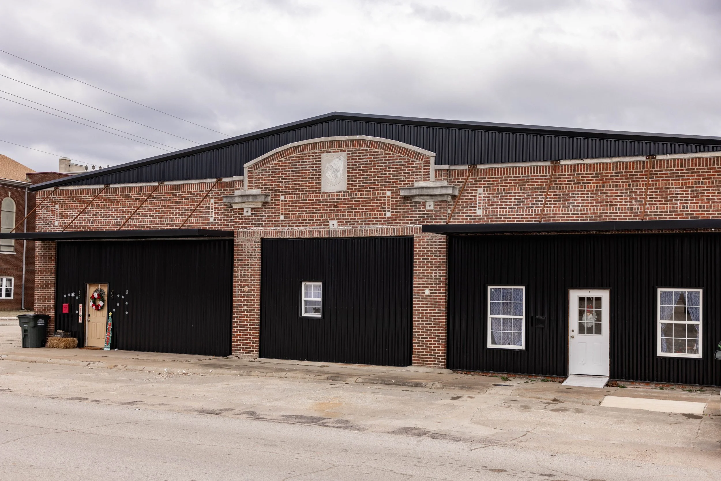 A building with brick and black siding, three doors, two windows, and Christmas decorations outside on a cloudy day.