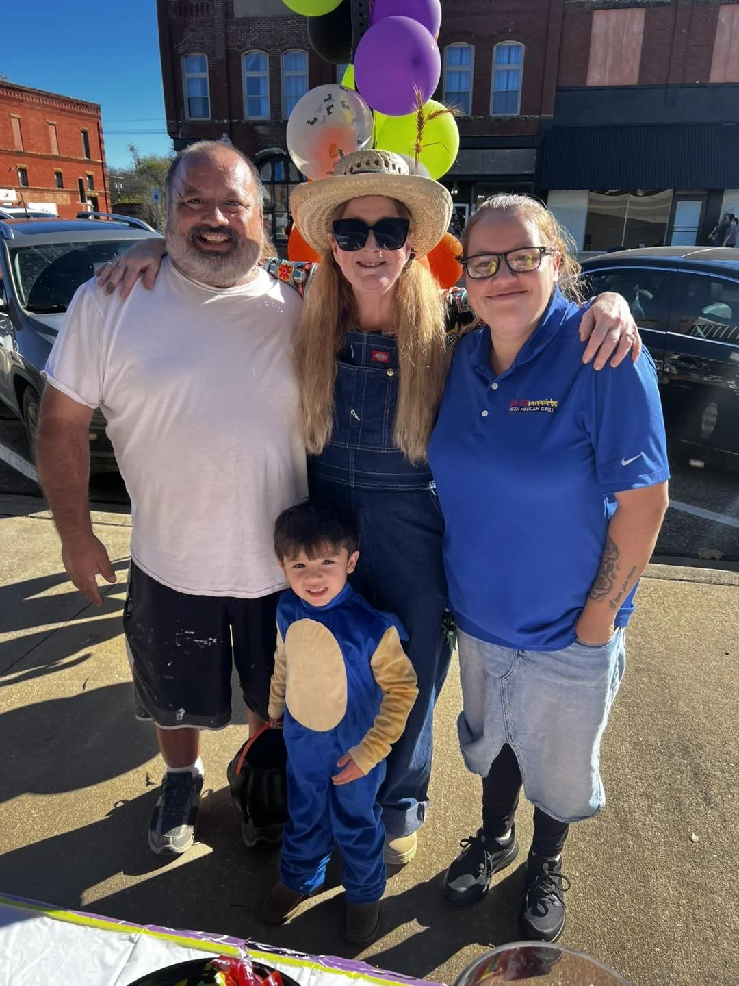 A group of four adults and one child smiling outdoors during a festive event, with balloons and a decorated table in the foreground.