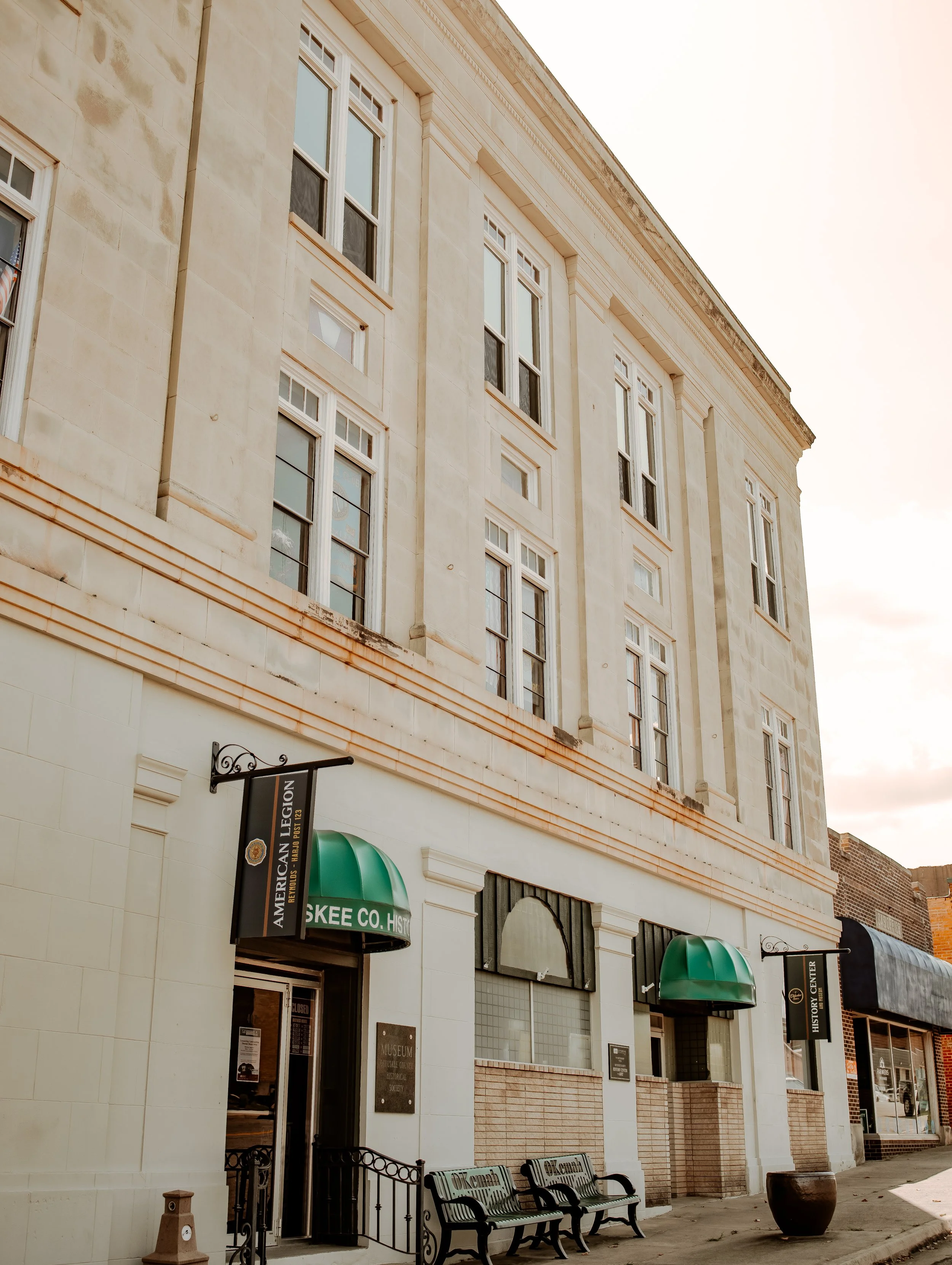 A multi-story beige building with green awnings over the windows and entrance, sign reading 'American Legion' and benches outside on the sidewalk.
