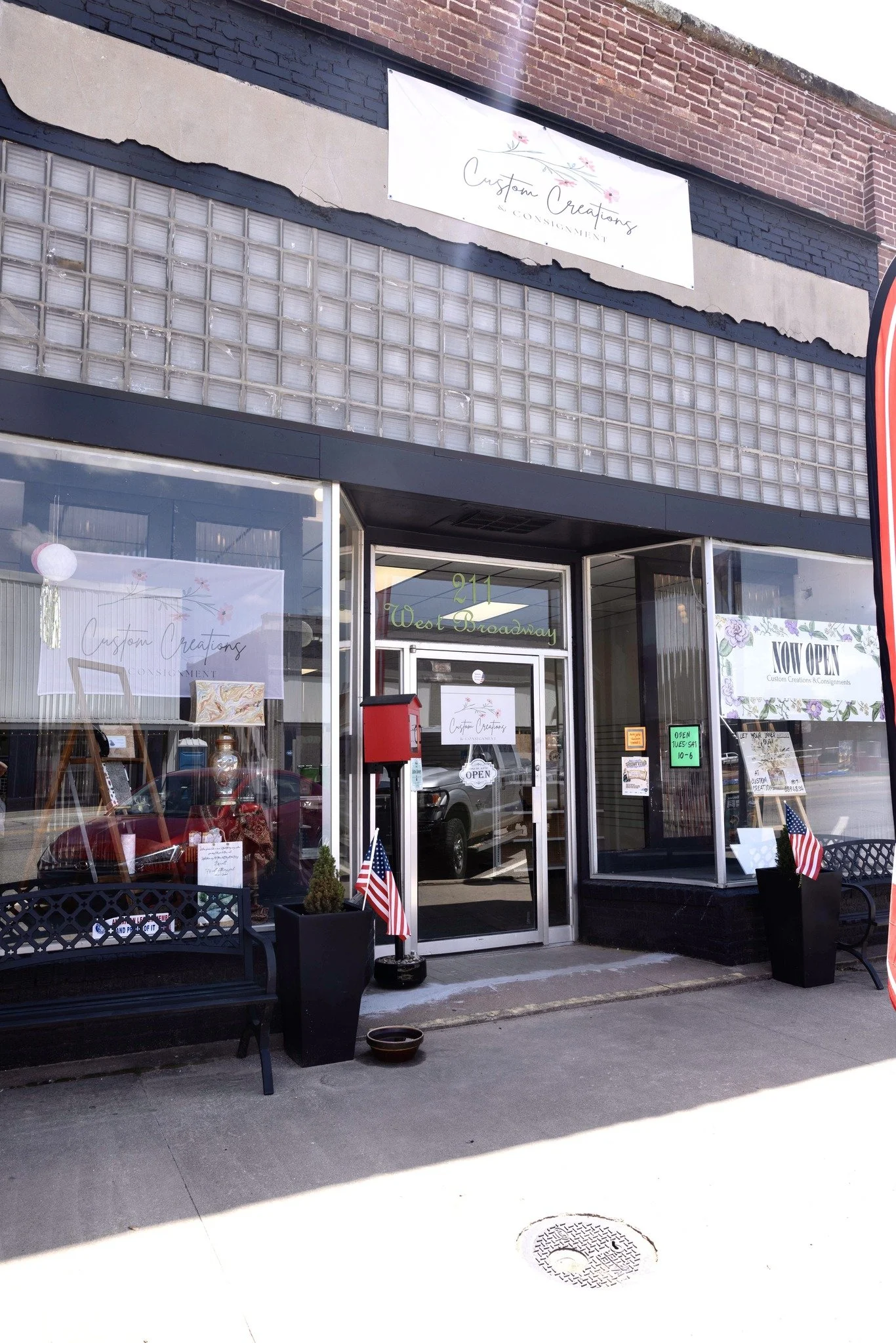The storefront of a shop named 'Custom Creations & Conciement' at 211 West Broadway, with two American flags and a bench outside. Signs indicate the shop is open.
