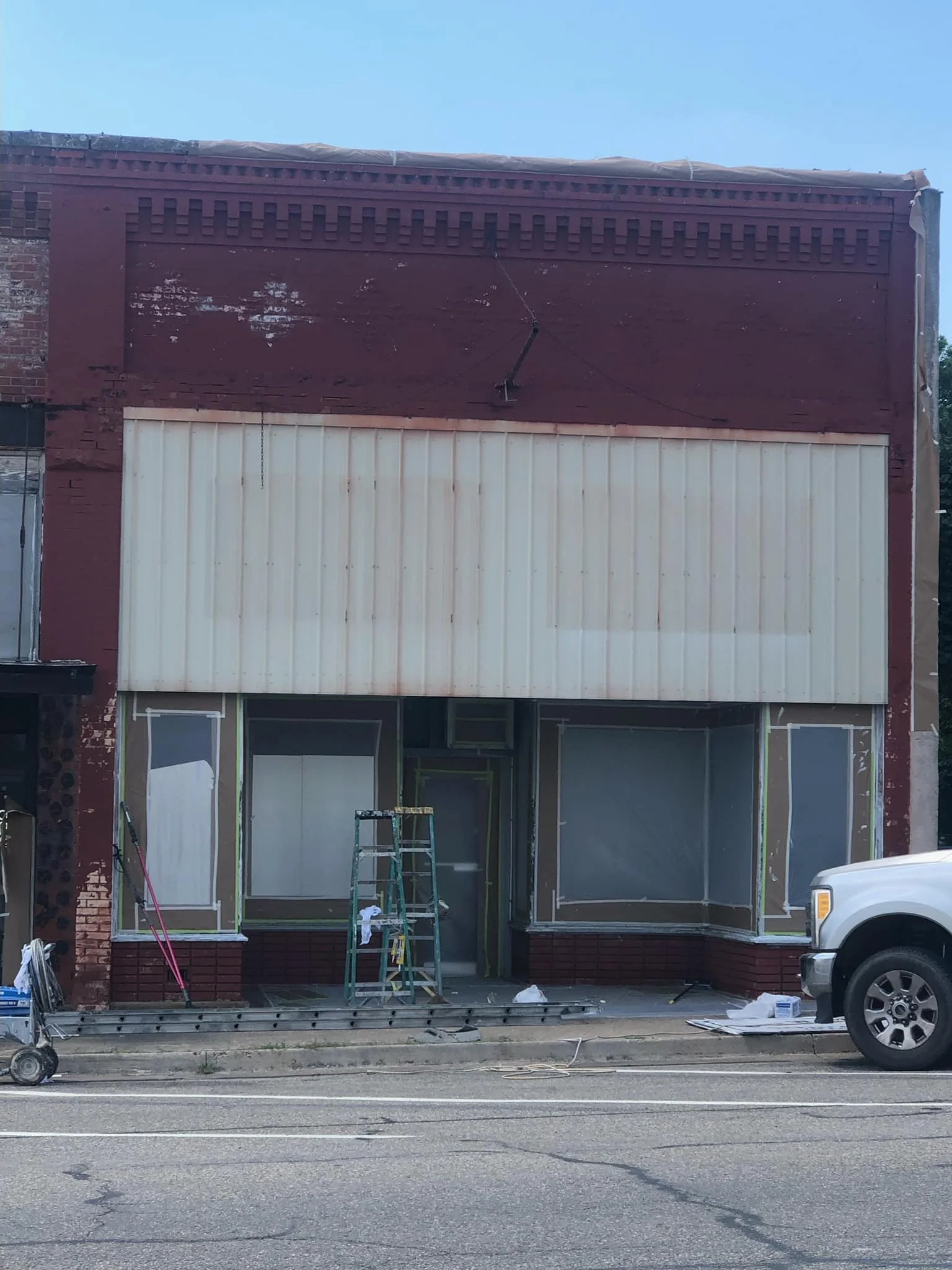 A building under renovation with windows covered in plastic, a ladder in front, and construction tools on the sidewalk, next to a parked white truck on the street.