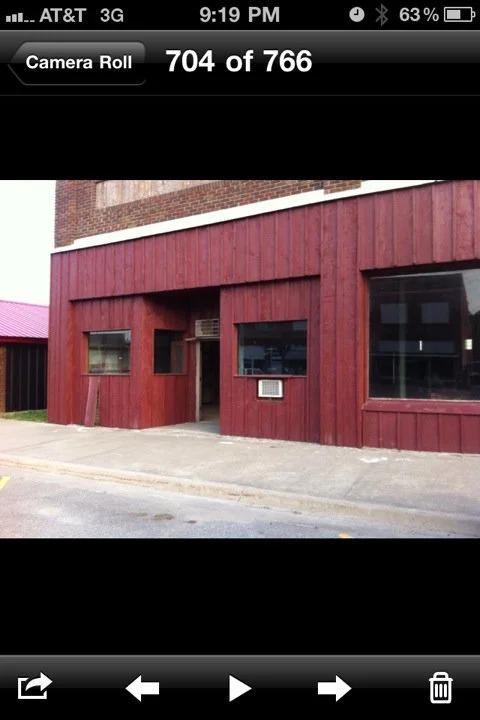 Red wooden building with two windows, a small door, and an air conditioning unit outside, located at the sidewalk near a street.