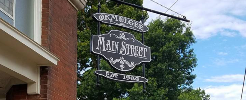 Black and white street sign for Main Street, established in 1986, hanging on a pole in front of green trees and a brick building.